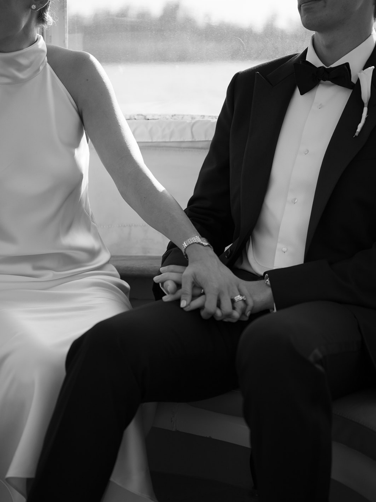 A black and white photo of a bride and groom holding hands, sitting next to each other, with the bride in a satin dress and the groom in a tuxedo with a bow tie.