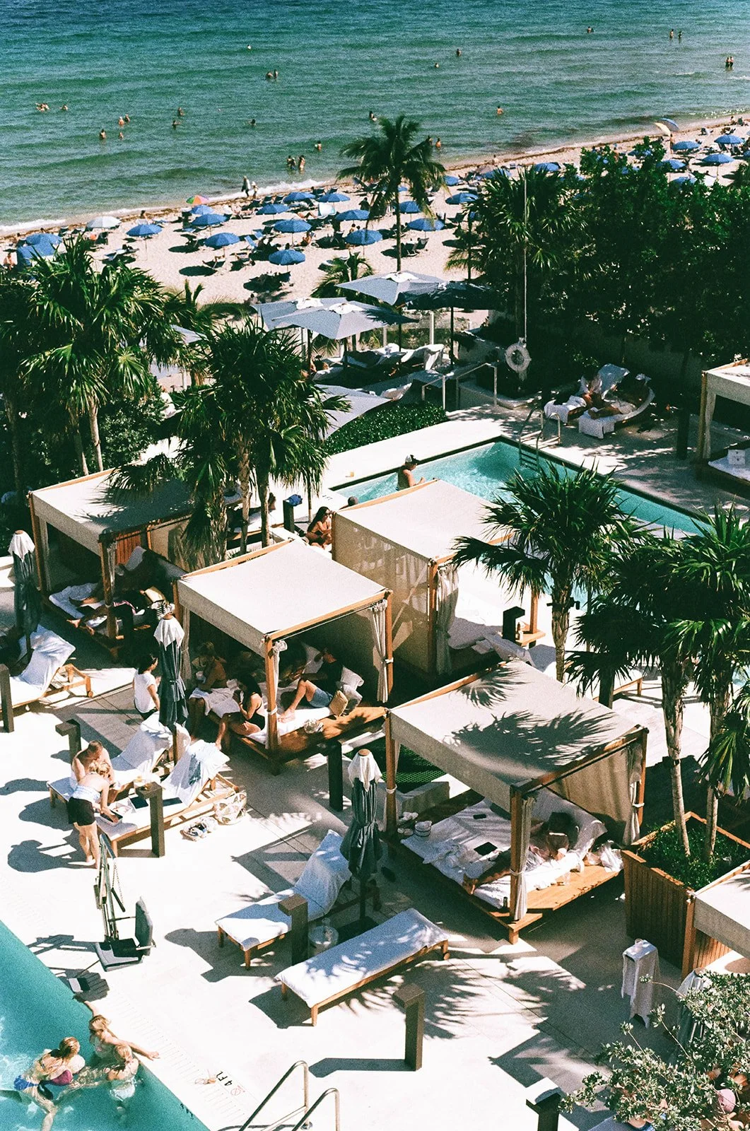 Aerial view of a beach resort with swimming pool and cabanas, palm trees, people relaxing on lounge chairs, and swimming in the ocean with umbrellas.