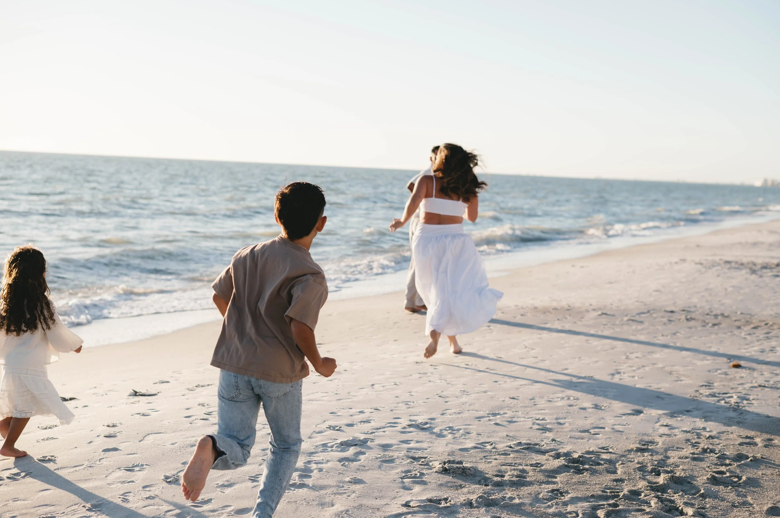 Group of people running and playing on a sandy beach near the ocean, with the sun shining brightly in the sky.