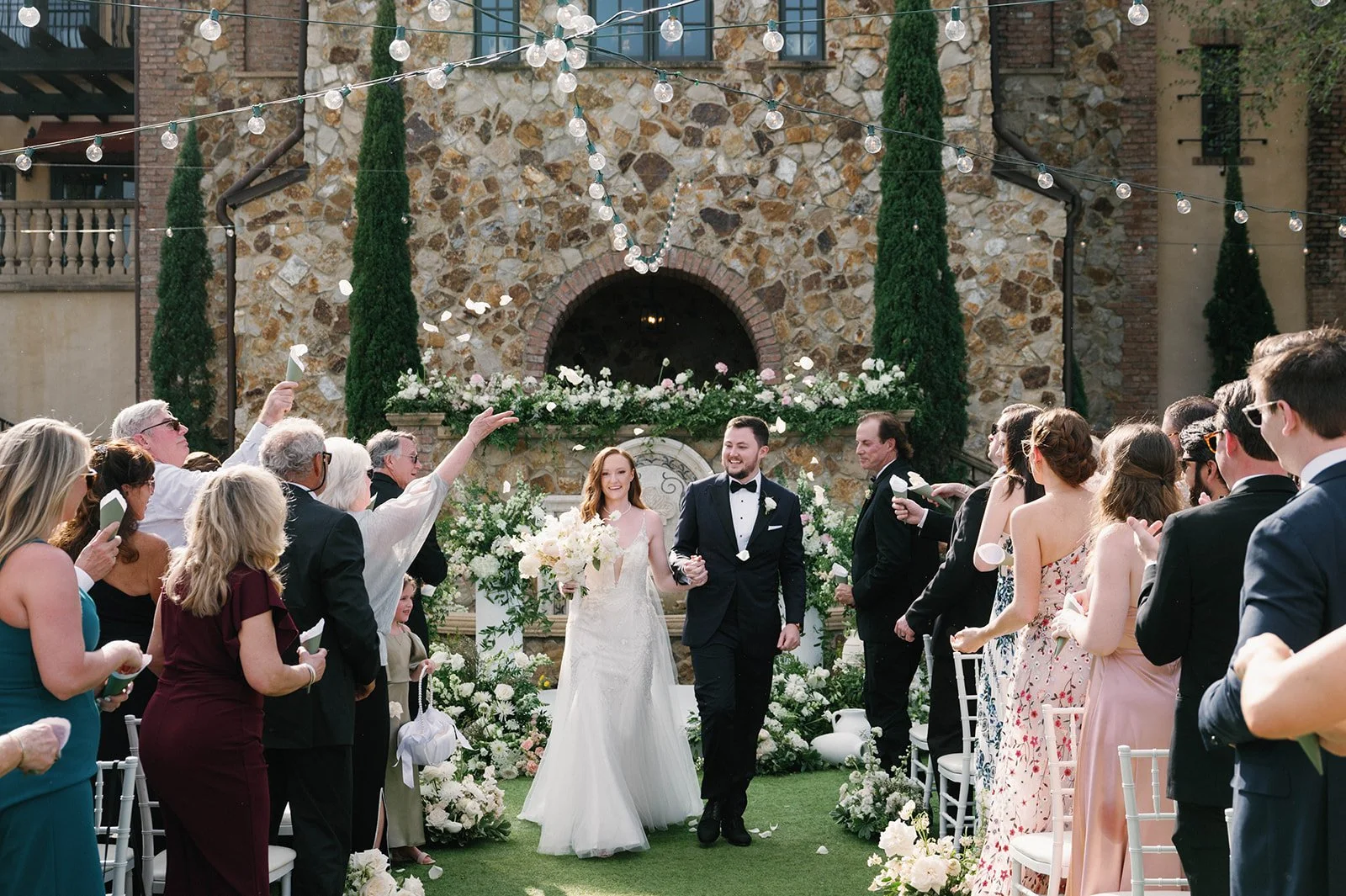 A wedding ceremony with a bride and groom walking hand in hand, surrounded by guests, flowers, and outdoor garden decor.