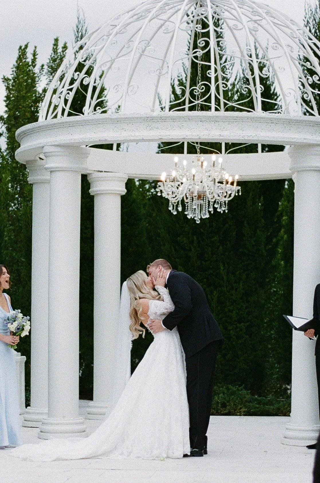 A wedding ceremony taking place outdoors under a white gazebo with columns, a chandelier, and a decorative metal dome. A bride and groom are kissing, with the bride in a white gown and the groom in a black suit. A bridesmaid holding a bouquet is visi