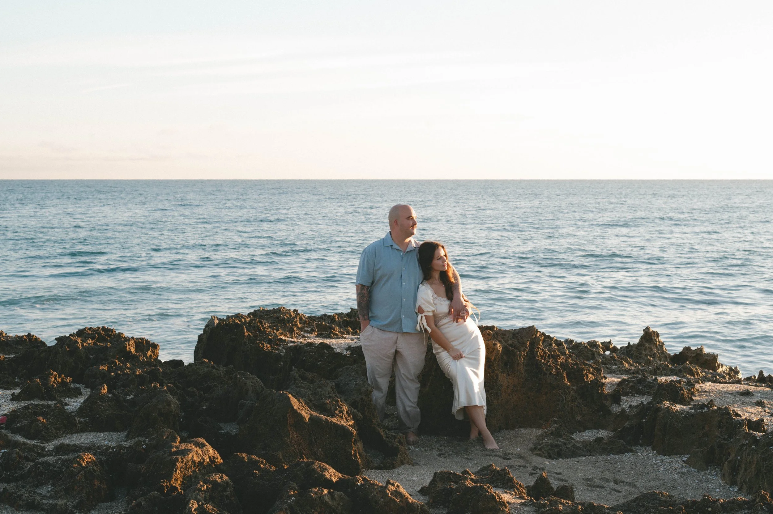A couple standing on rocky shoreline by the ocean during sunset, with the man in a light blue shirt and the woman in a cream dress, both looking out at the water.