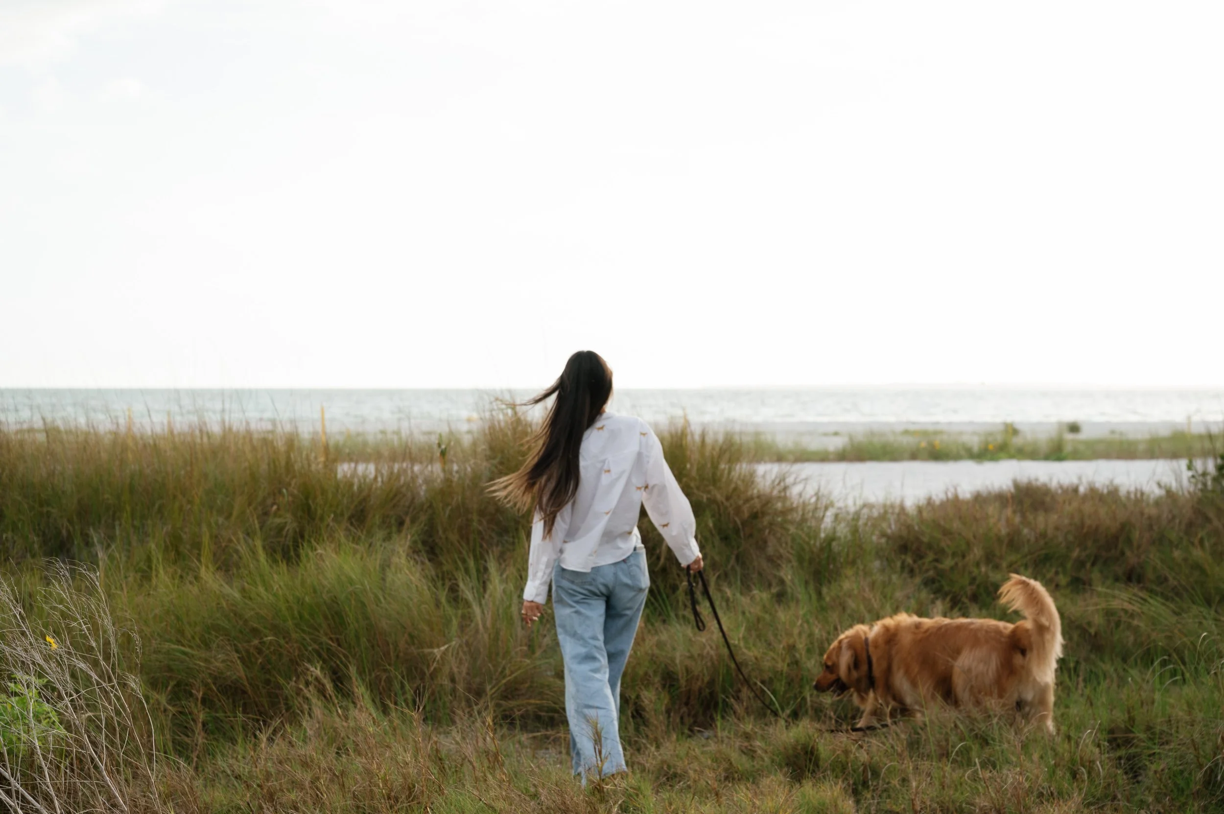 A person with long brown hair, wearing a white shirt and light blue jeans, is walking a dog along a grassy path near the beach, with the ocean in the background.