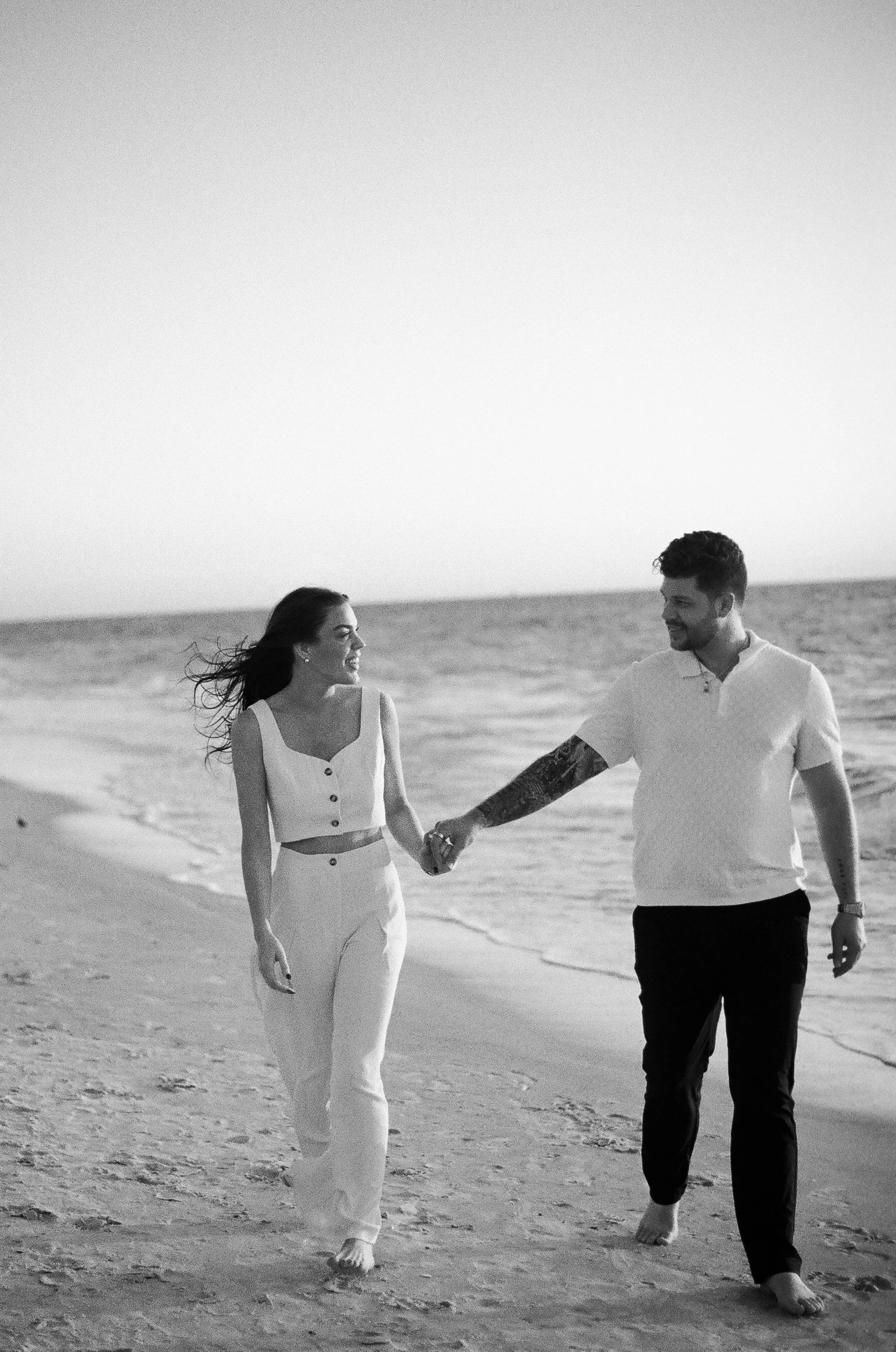 A black-and-white photograph of a man and woman walking hand-in-hand along the beach, smiling at each other.