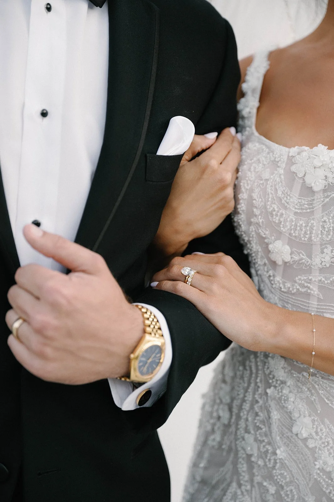 Close-up of a bride and groom holding arms, the bride showing a wedding dress and ring, the groom in a black tuxedo with a white shirt, and gold watch.