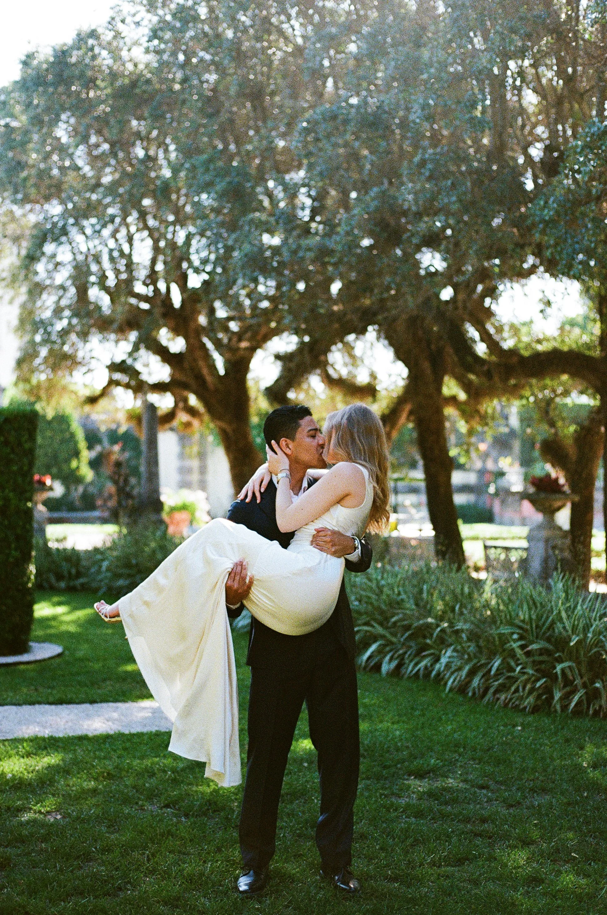 A man in a tuxedo holding a woman in a long dress in a park.