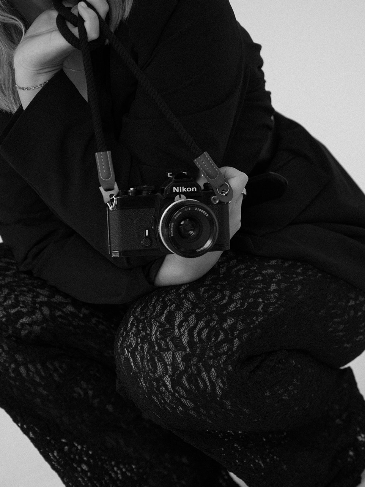 Black and white photograph of a person sitting cross-legged, holding a Nikon camera with a strap around their neck. The person wears a dark jacket, lace-patterned pants, and a bracelet on their wrist.