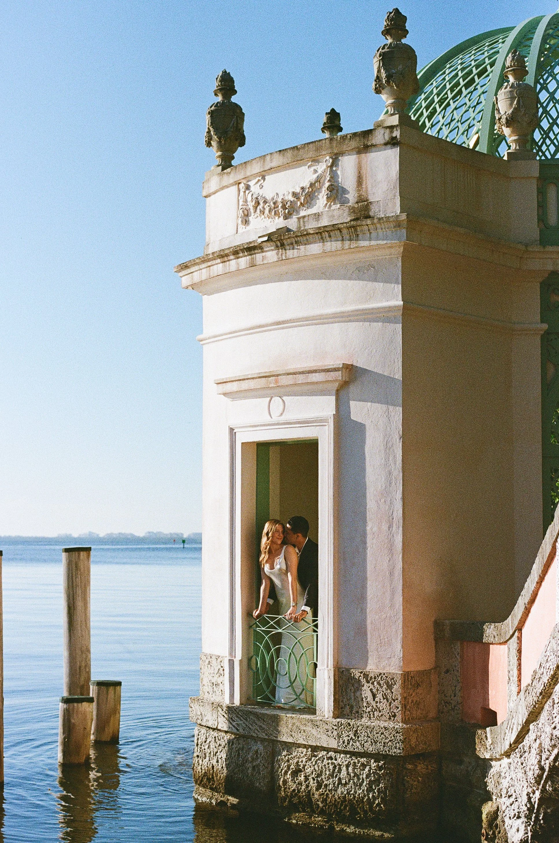 A couple in wedding attire standing on a balcony of a small building by the water, with a scenic view of a calm lake and distant shoreline in the background.