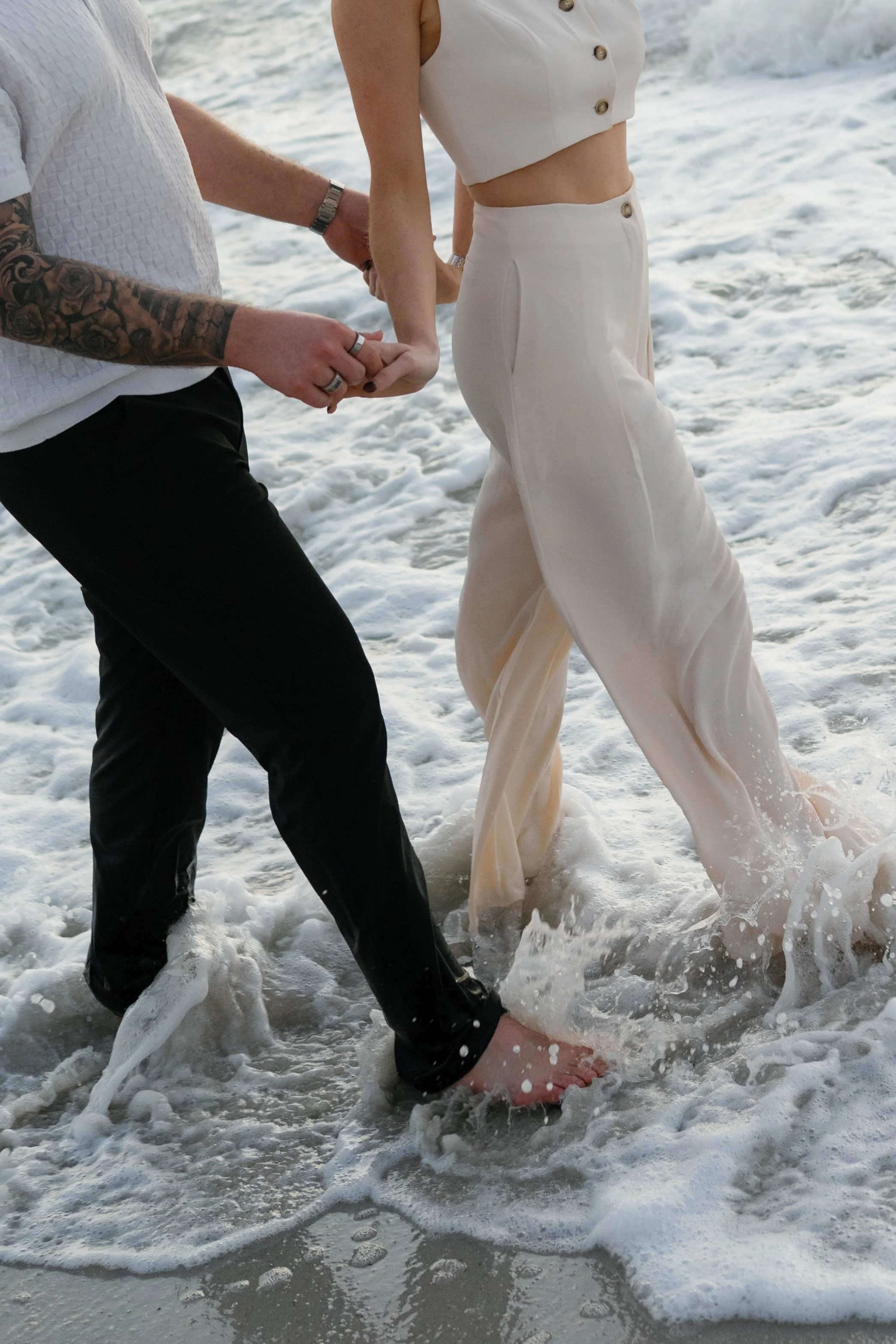 A couple holding hands in the water at the beach, with their feet in the waves, seen from the waist down.