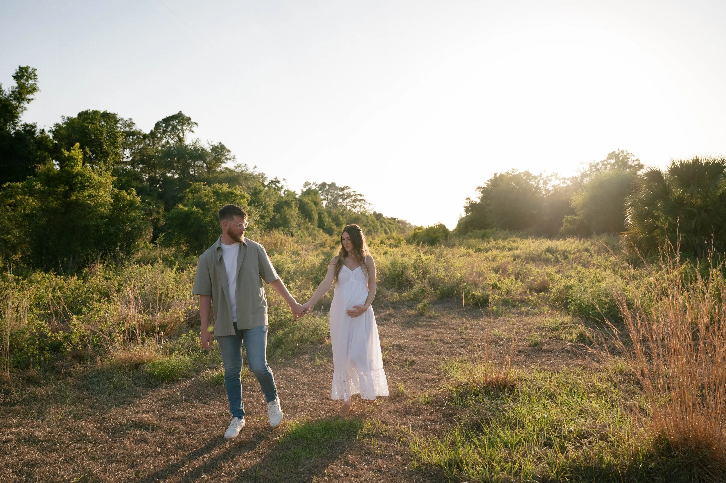 A couple walking hand-in-hand outdoors in a grassy field during sunset, with trees and clear sky in the background.