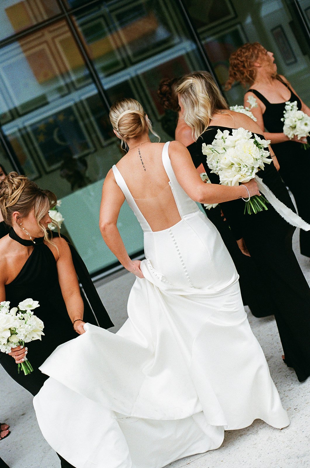 Women in formal black dresses holding bouquets of white flowers, one woman in a white wedding dress with an open back, is being assisted by the others, in front of a building with large glass windows.