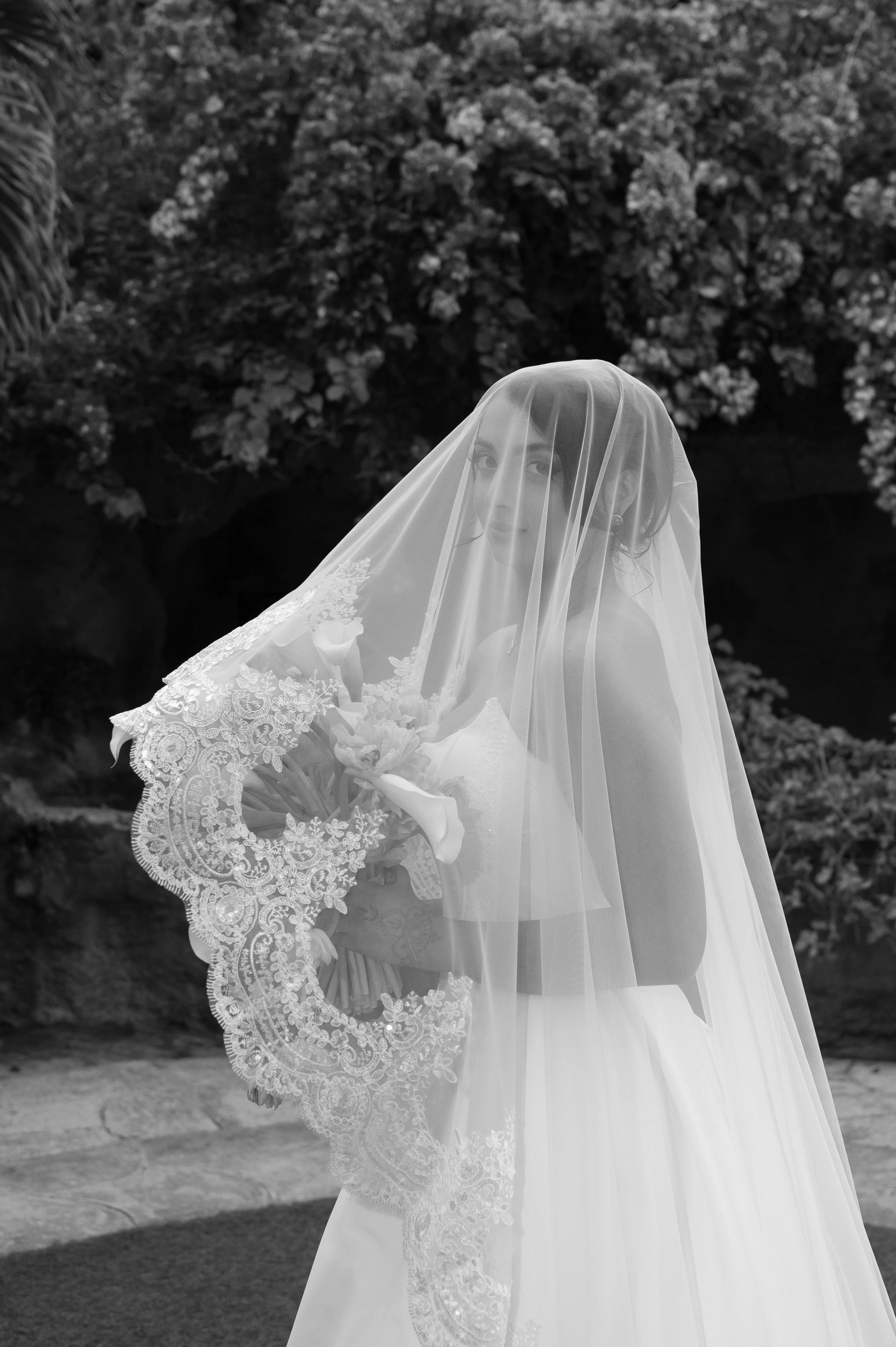 A bride wearing a lace wedding dress and a veil holding a bouquet of flowers outside in front of bushes.