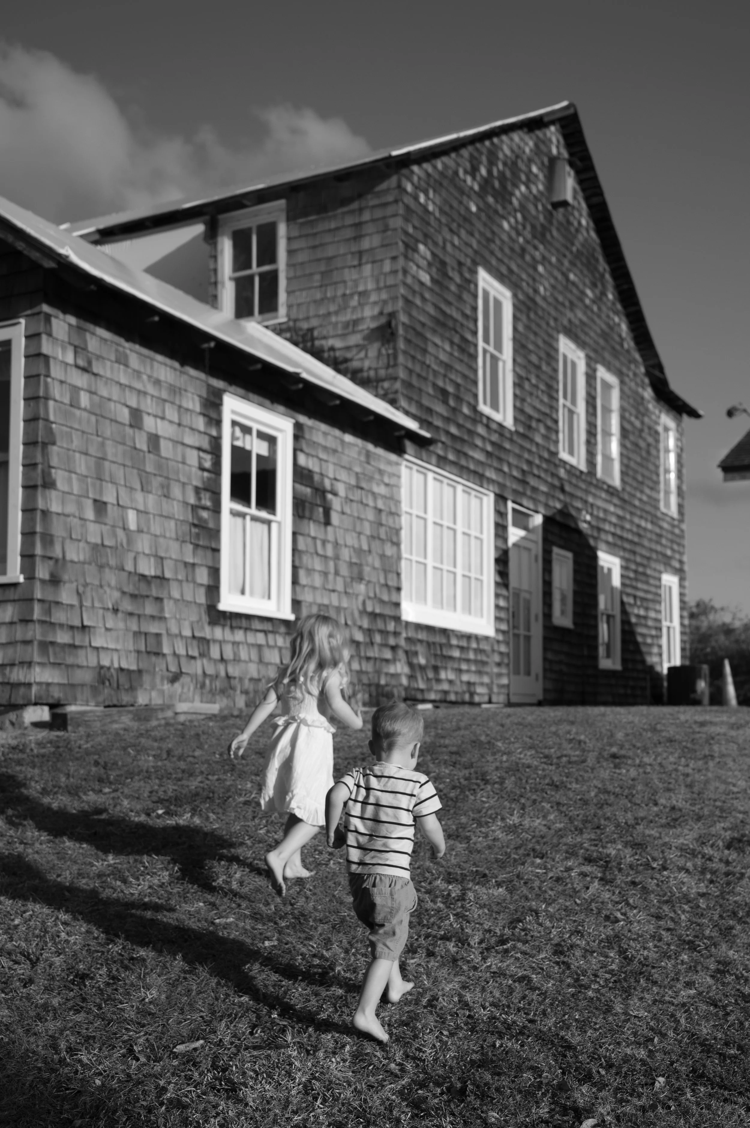 Two children running outside on a grassy area in front of a wooden house with multiple windows, in a black and white photo.