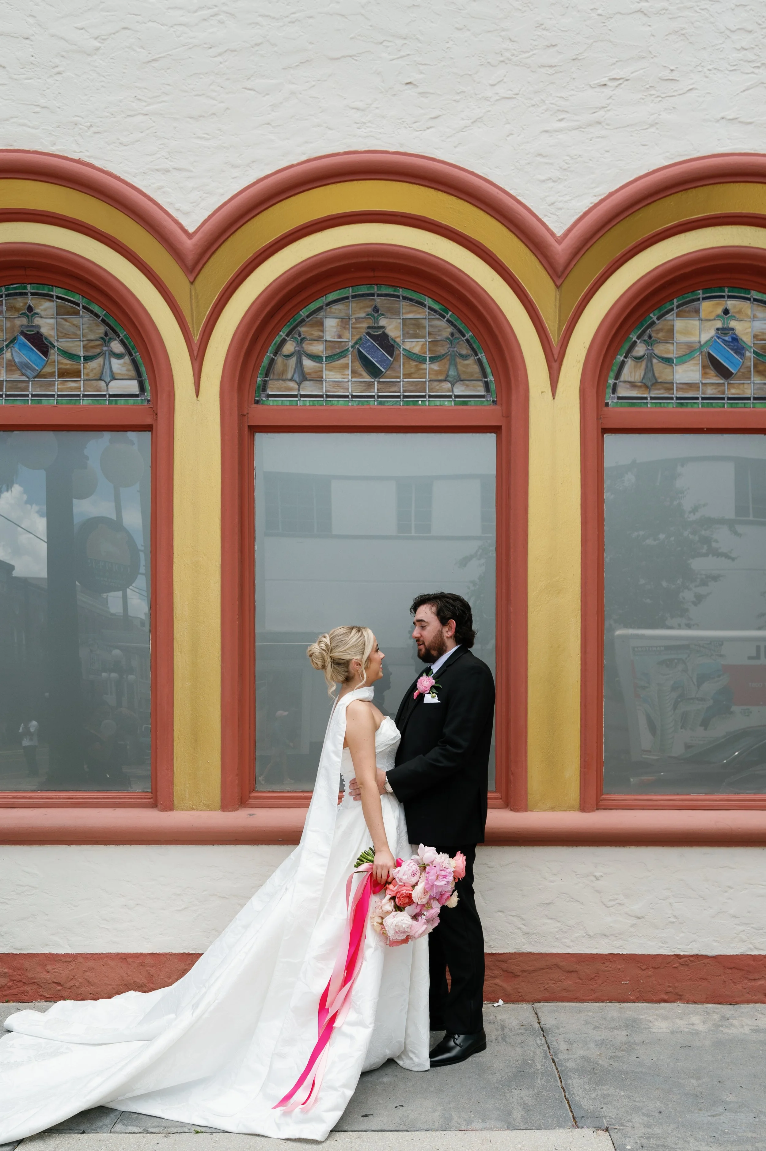 A bride and groom standing face to face in front of a colorful arched window, holding hands, with the bride holding a bouquet of pink and white flowers.