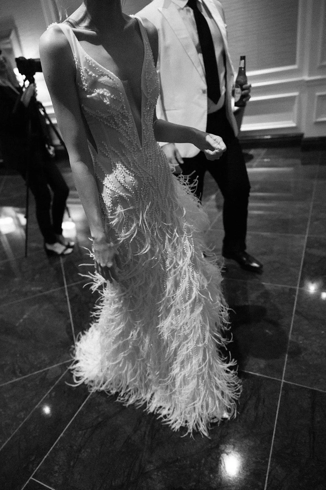Black and white photo of a woman in a fancy dress with feather details, standing next to a man in a shirt and tie holding a beer bottle at an indoor event.