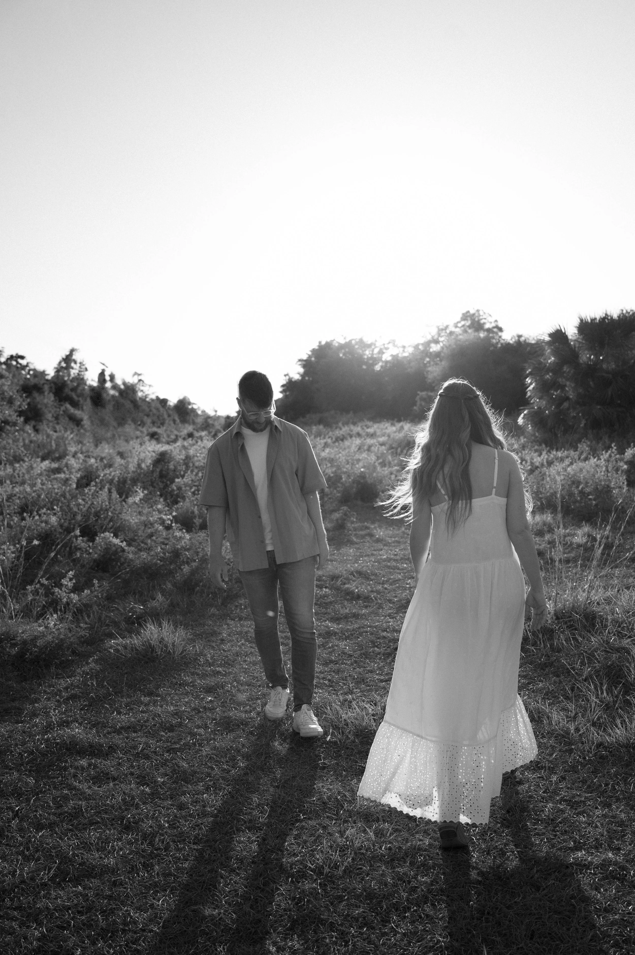 A black and white photo of a man and woman walking through a grassy outdoor area. The woman is wearing a long white dress, and the man is dressed casually in a shirt and jeans. The sunlight is shining brightly from behind, creating elongated shadows 