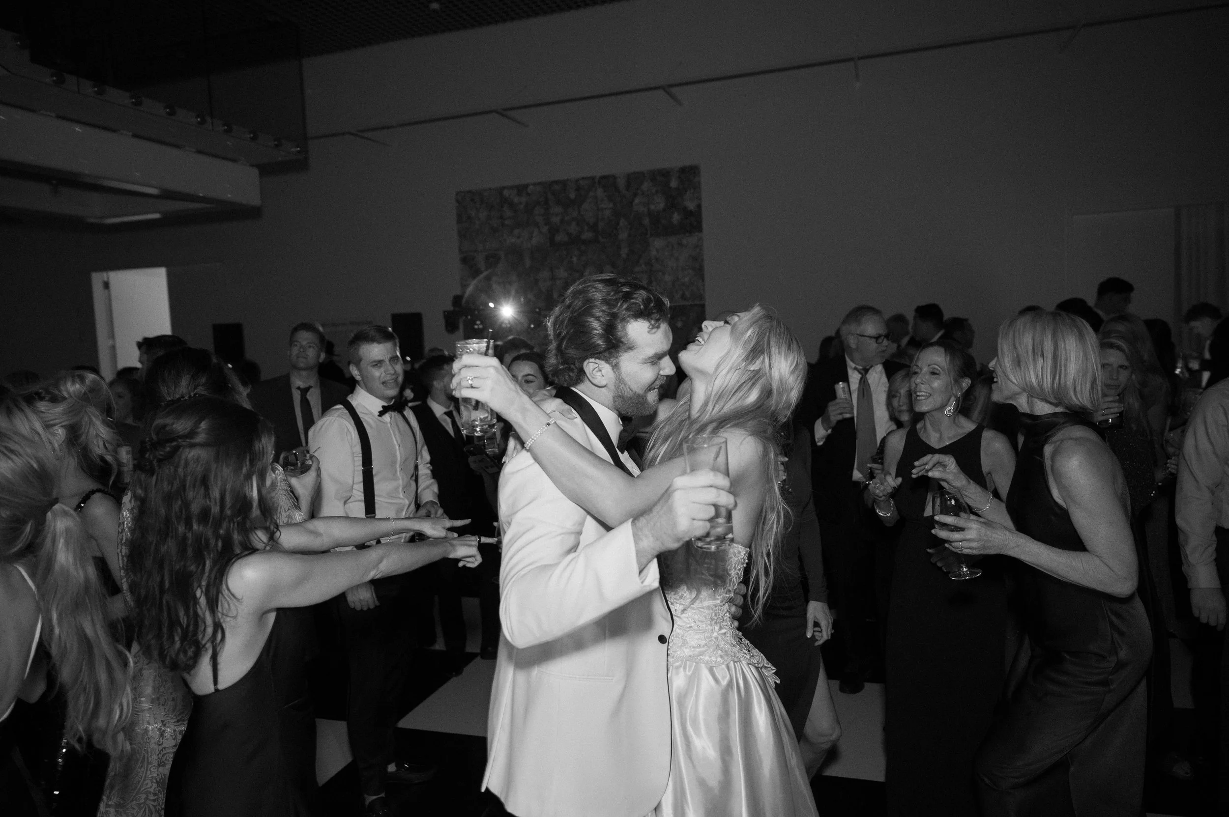 A black and white photo of a social gathering with people dancing and holding drinks. Two people in the foreground, a man and a woman, are embracing and smiling at each other.