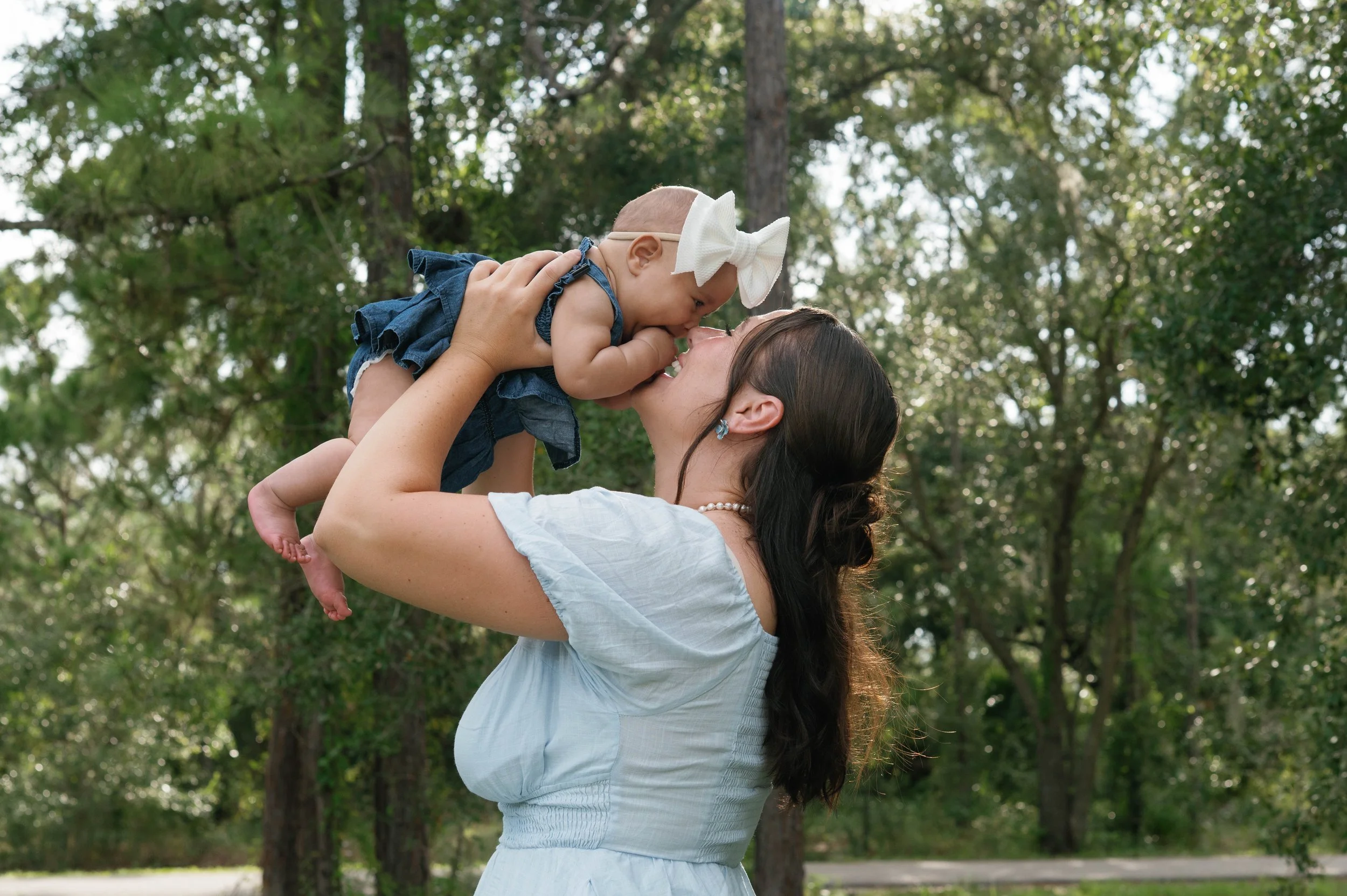 Woman lifting a baby girl outdoors among trees, both smiling, woman wearing a white blouse, baby in a dark blue dress with a large bow headband.