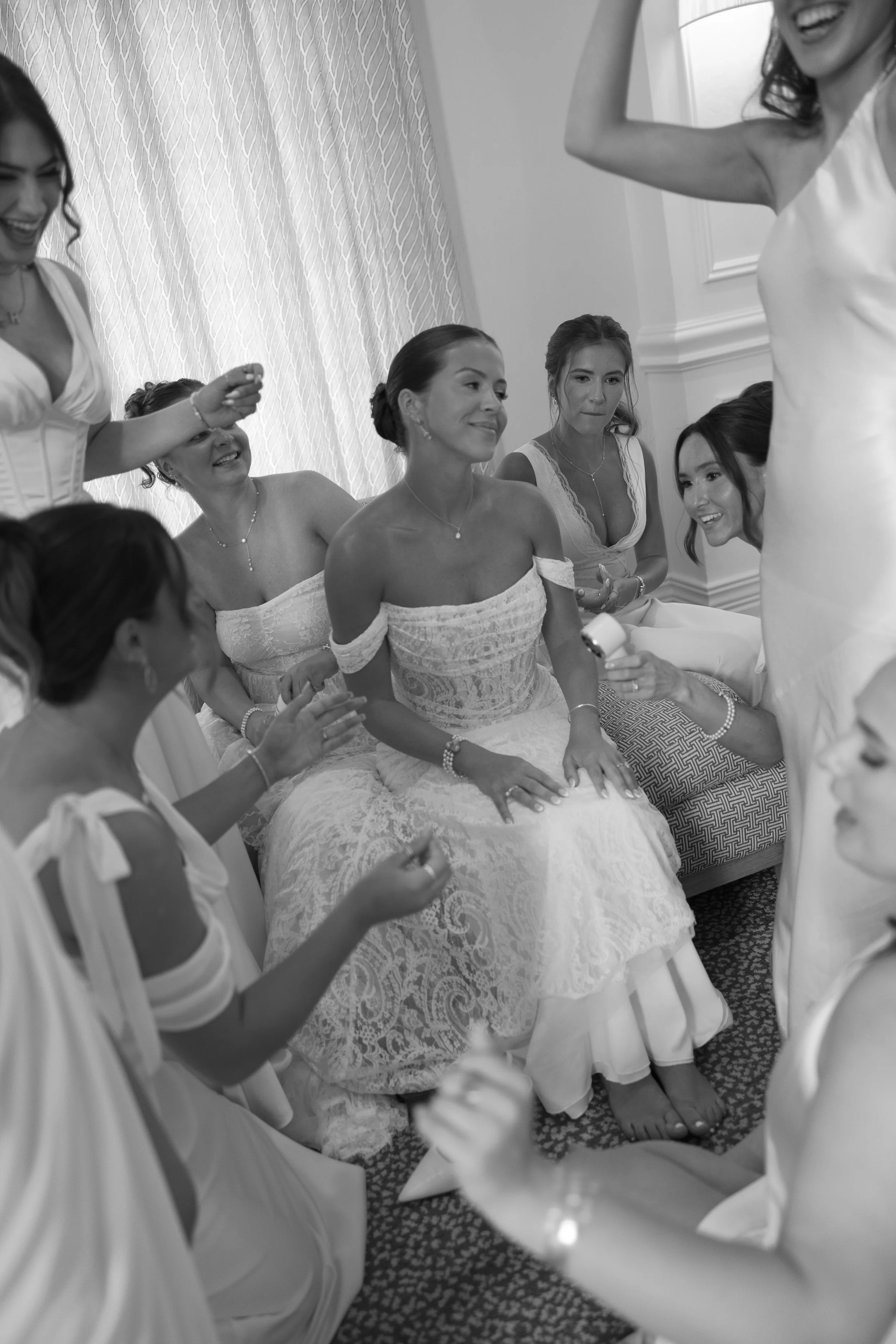 A bride sitting on a chair surrounded by women in wedding dresses, smiling and laughing during a wedding preparation in a well-lit room.