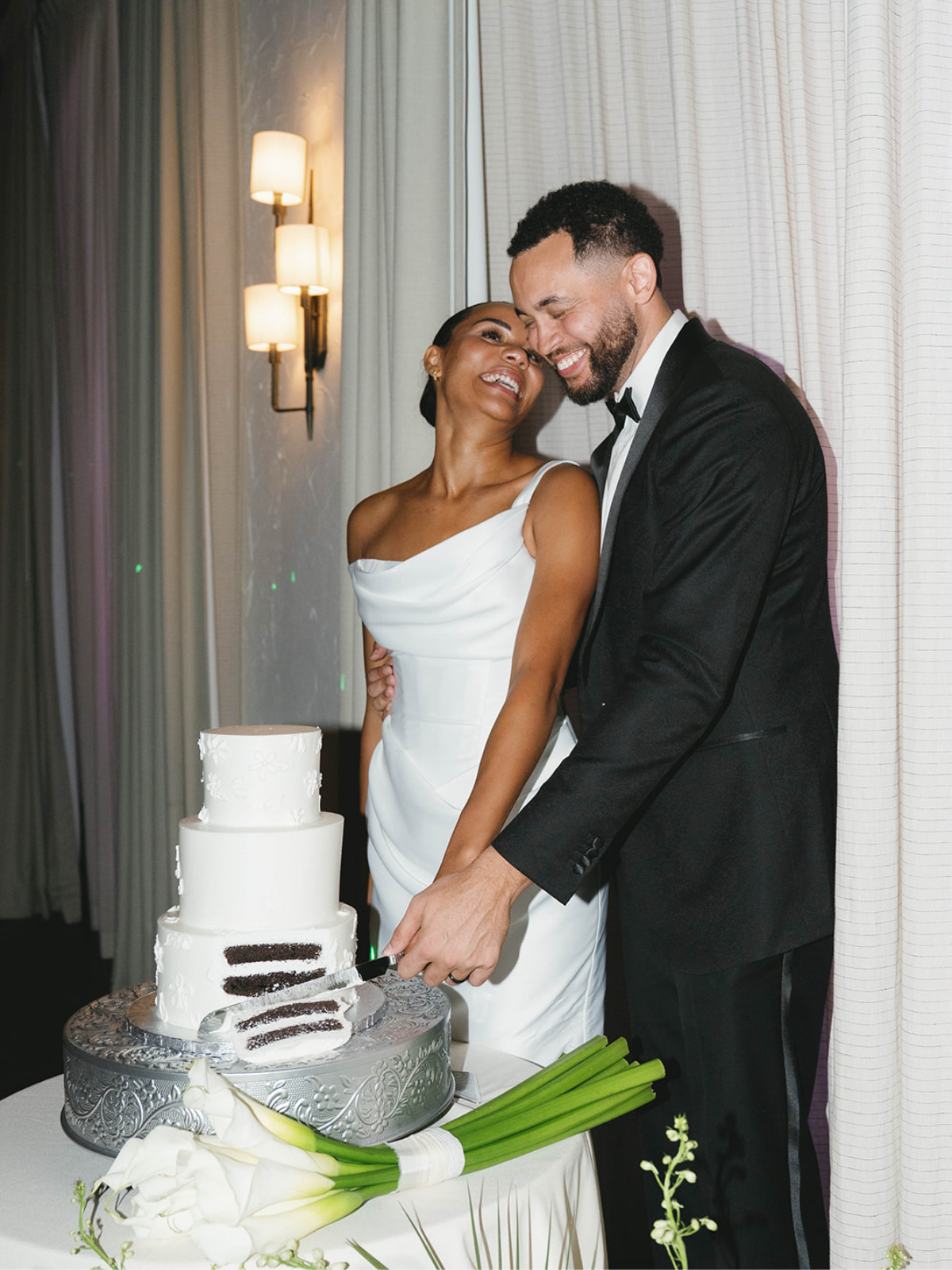 Married couple in wedding attire cutting a wedding cake together at their reception, with soft lighting and floral decorations nearby.
