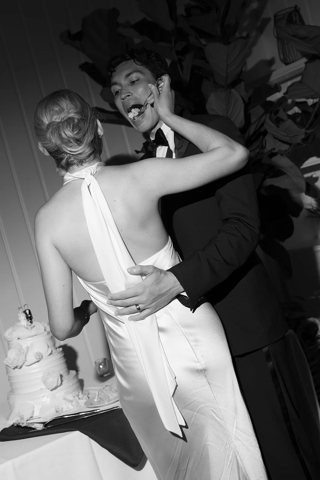 A black-and-white photo of a bride and groom dancing closely at their wedding reception. The bride is wearing a sleeveless satin dress, and the groom is in a tuxedo. The bride is feeding cake to the groom with a fork. There is a wedding cake in the b