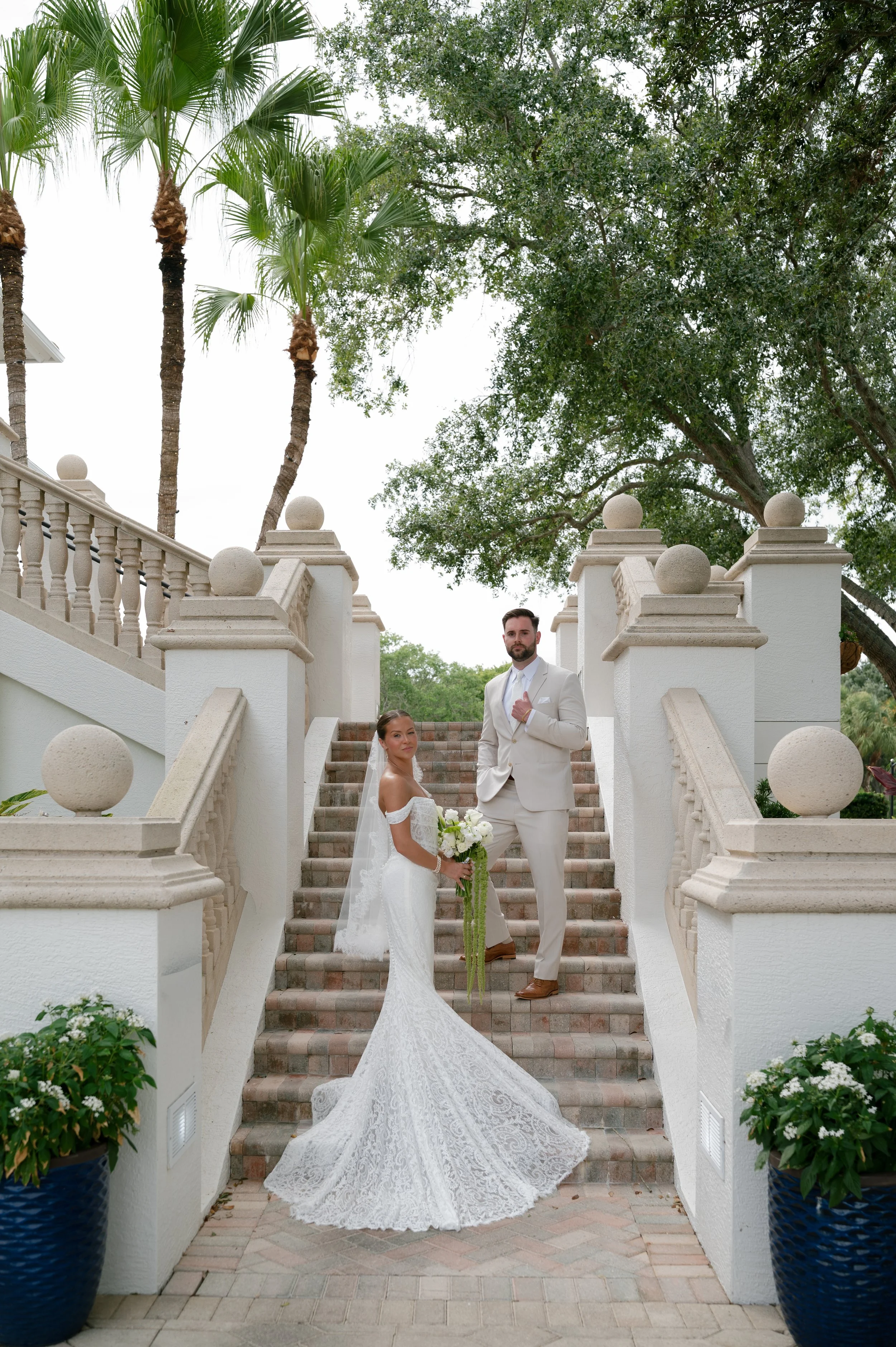 A bride and groom standing on outdoor staircase with ornate white railing, surrounded by greenery and tall palm trees, during a wedding photoshoot.