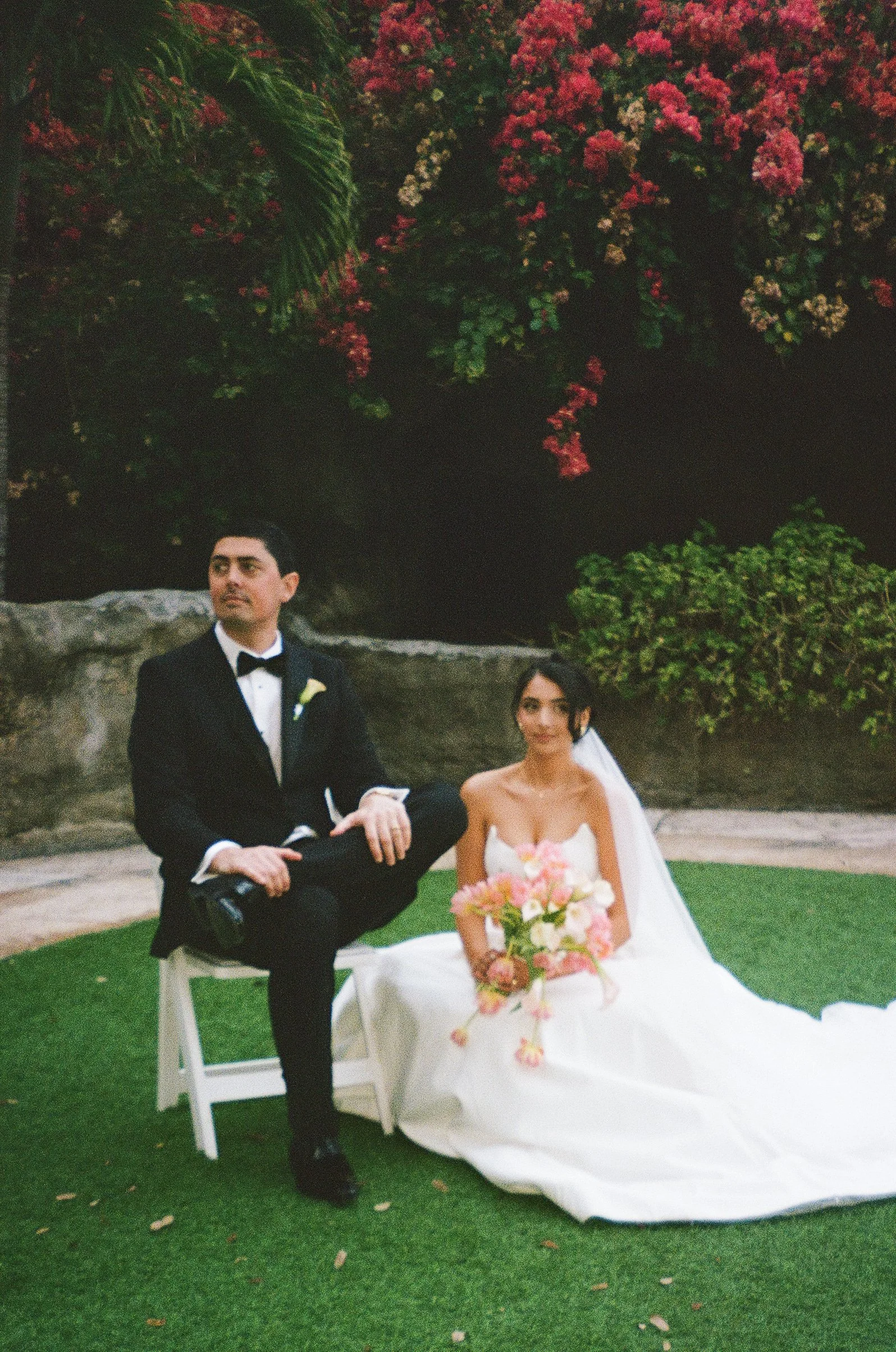A bride and groom at their wedding, with the groom in a tuxedo and the bride in a strapless white wedding gown holding a bouquet of pink and white flowers, seated outdoors on a green lawn with flowers and bushes in the background.