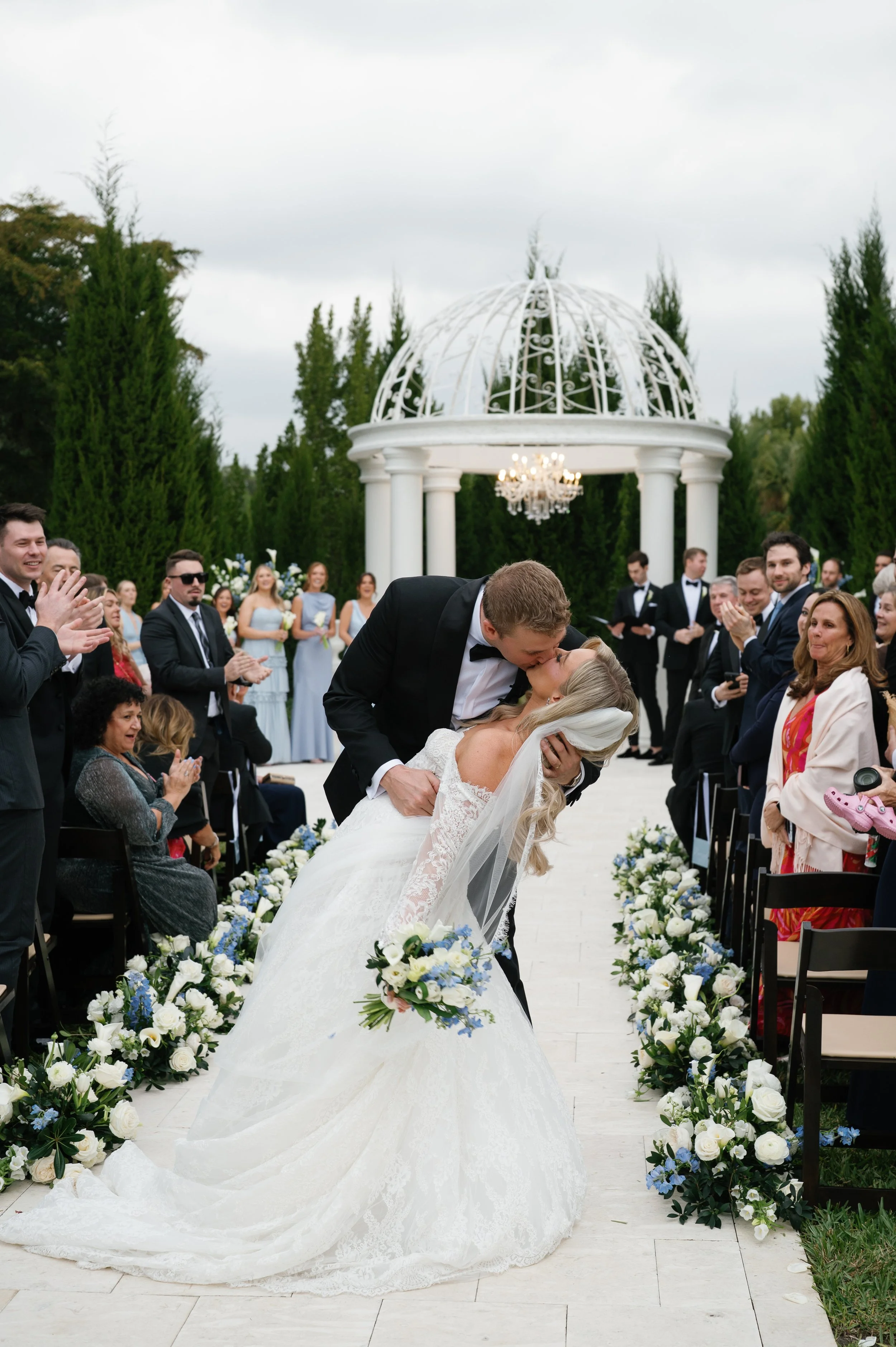 A wedding ceremony with a bride and groom kissing in front of guests outdoors, decorated with white and blue flowers along the aisle, near a white gazebo and surrounded by green trees.