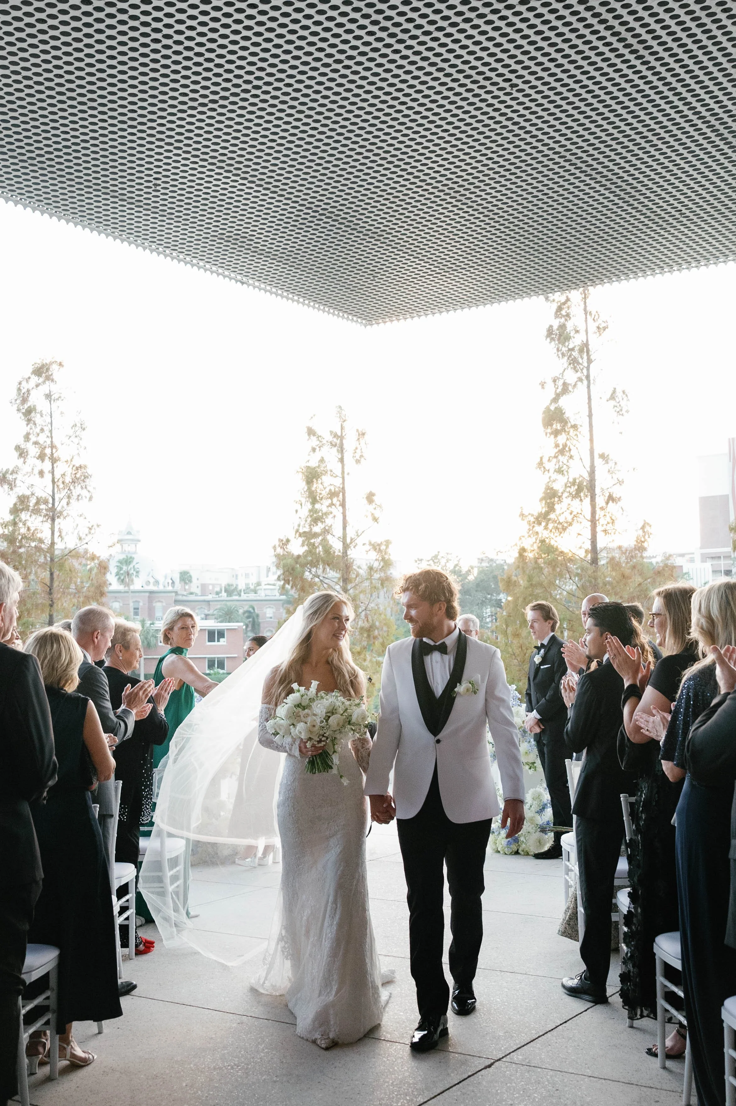A bride and groom holding hands and walking down the aisle at their wedding, surrounded by guests clapping and celebrating indoors with large windows and trees visible outside.