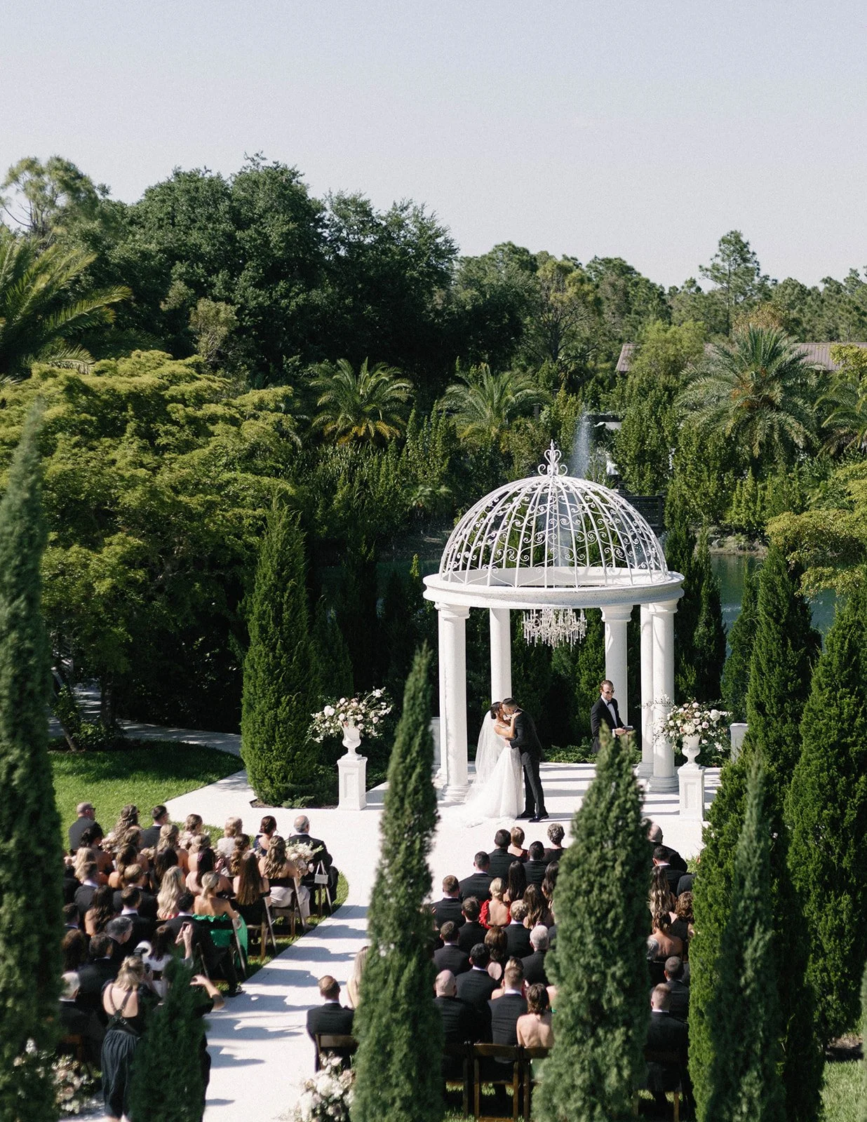 A wedding ceremony taking place outdoors in a lush garden with a white gazebo, where a bride and groom are sharing a kiss. Guests are seated in front, and the scene is surrounded by tall green trees and landscaped greenery.
