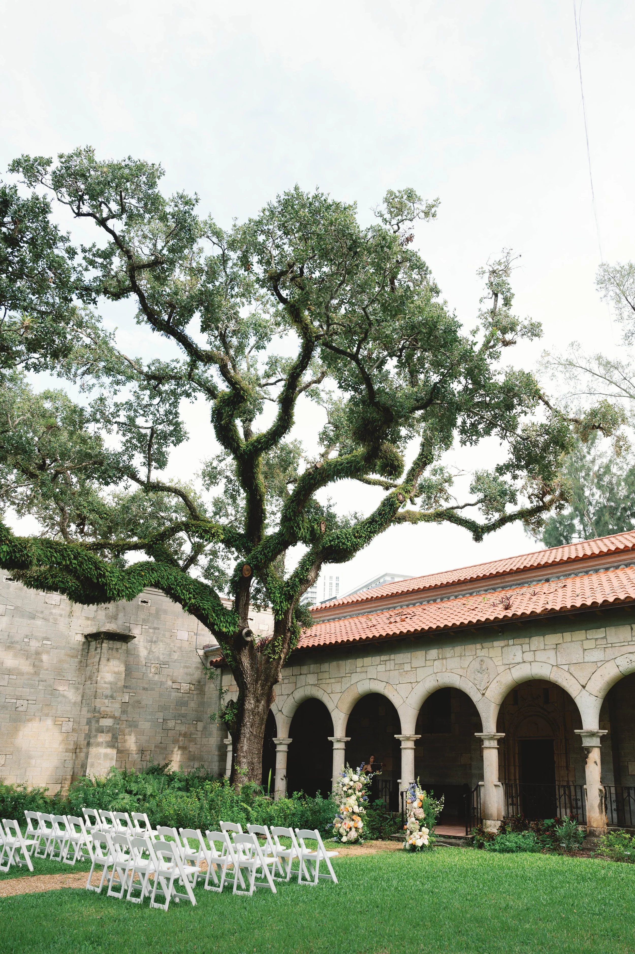 Outdoor wedding ceremony setup with white chairs arranged in front of an old stone building with arches, decorated with floral arrangements, beneath a large tree with lush green leaves.