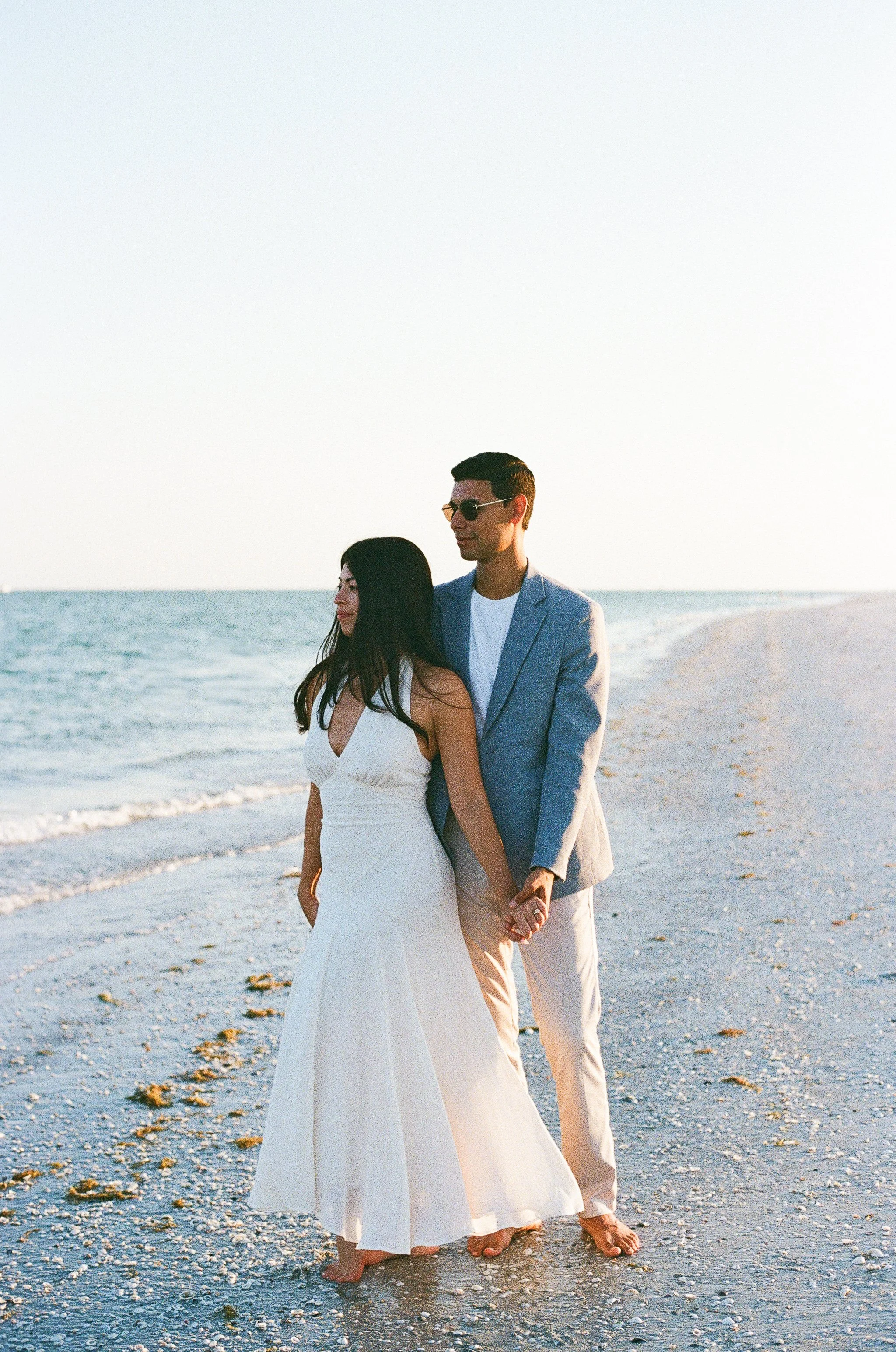A couple holding hands and walking on the beach during sunset, the woman in a white dress and the man in a light gray blazer and sunglasses.