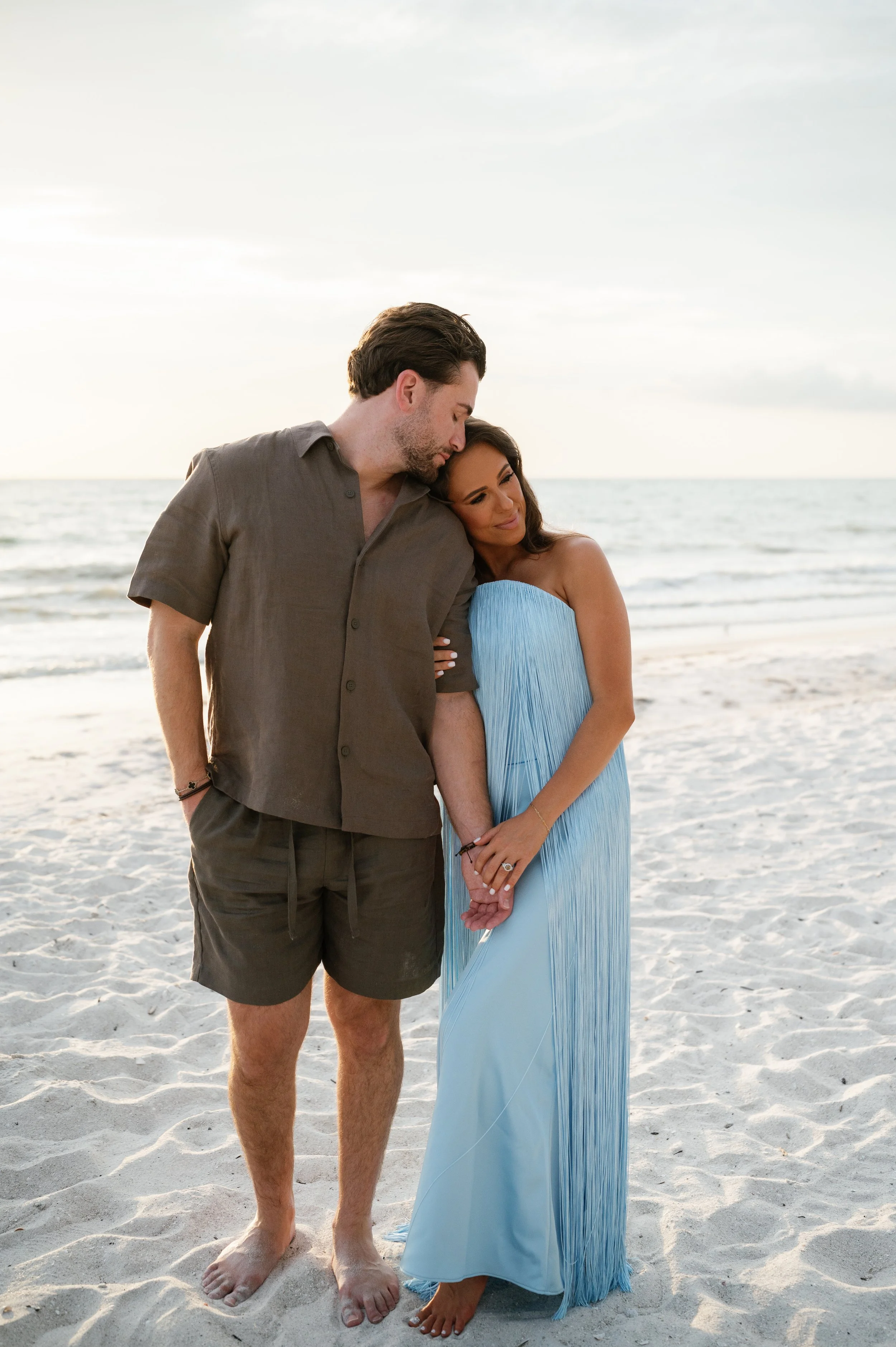 A couple holding hands and leaning their heads against each other on the beach, with the ocean and sky in the background during sunset.