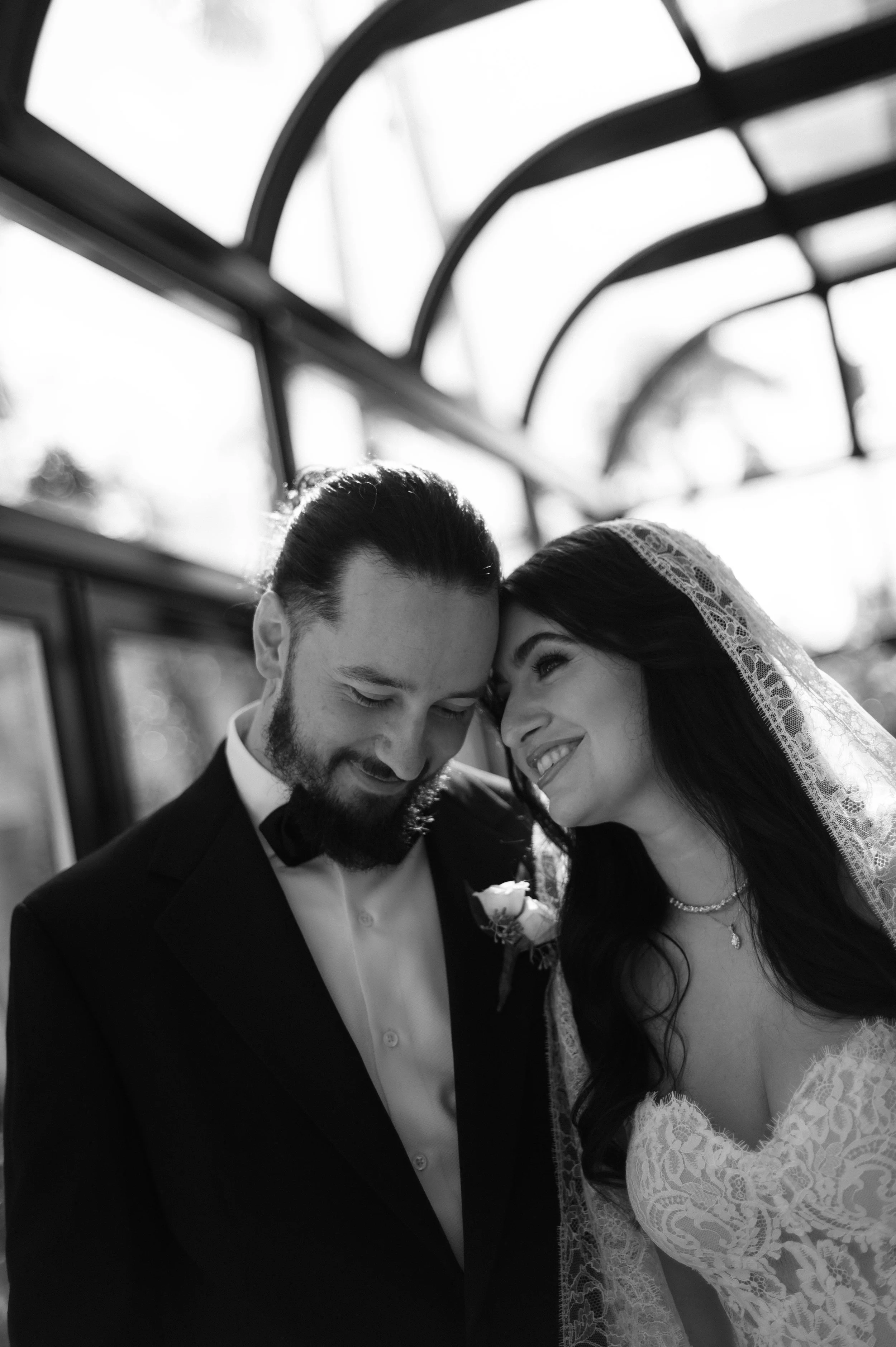 A black and white photograph of a bride and groom on their wedding day, standing close with foreheads touching, smiling lovingly at each other inside a vintage tram car.