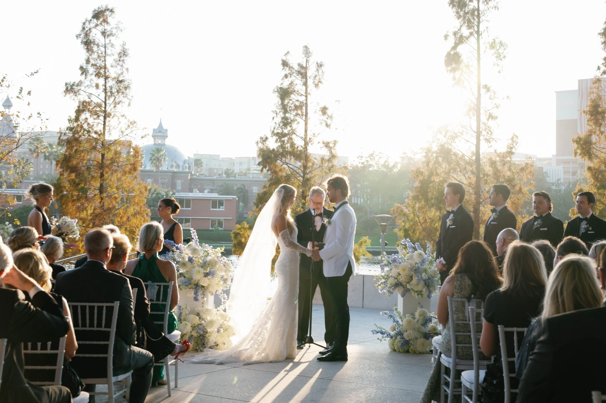 A wedding ceremony taking place outdoors at sunset with a bride and groom exchanging vows, surrounded by guests seated on white chairs, with trees and a cityscape with historic buildings in the background.