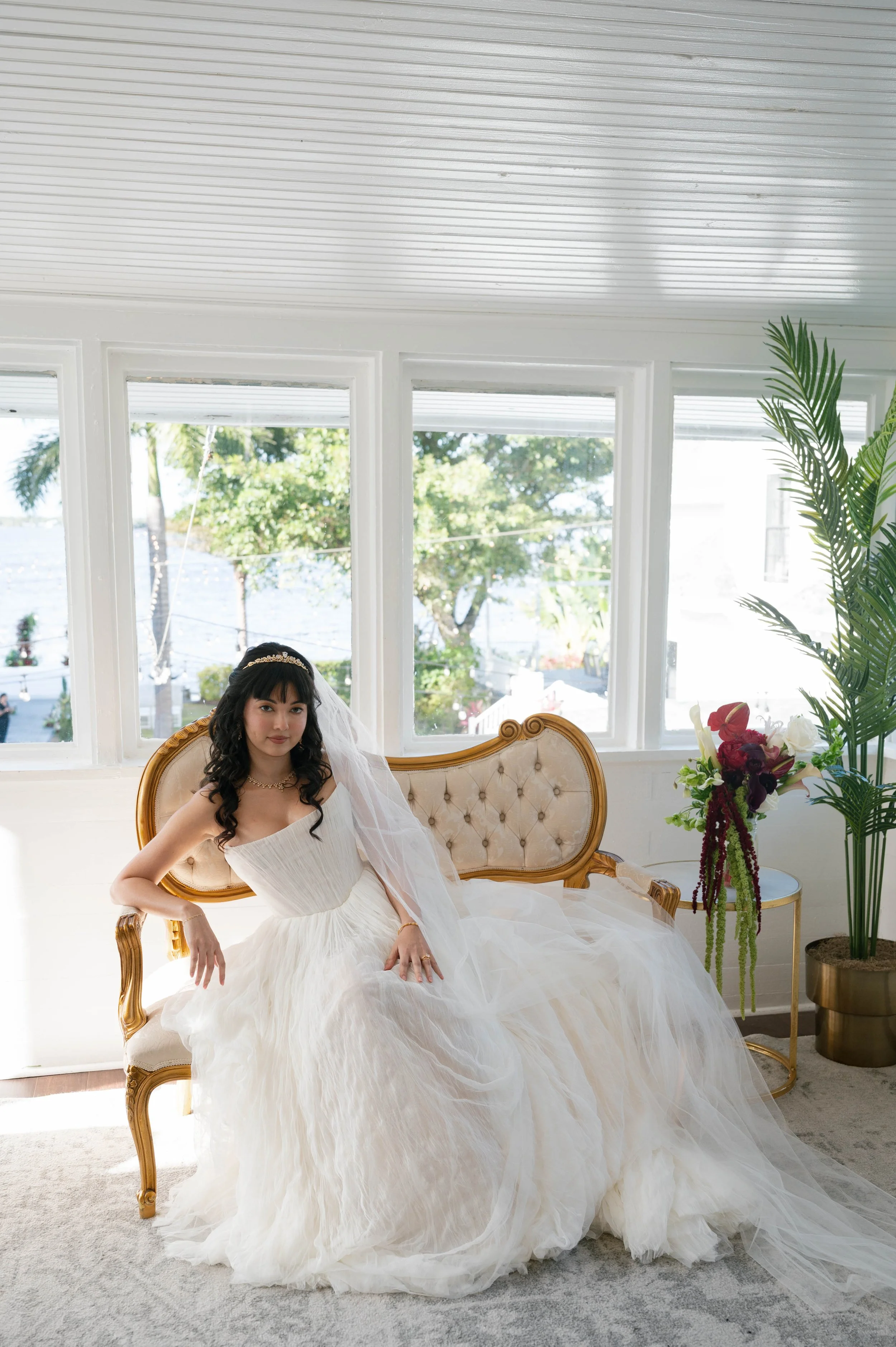 A woman dressed in a white wedding gown sitting on a vintage tufted sofa with a gold frame, inside a bright room with large windows and a view of trees and water outside. There is a tall green plant and a bouquet of flowers nearby.