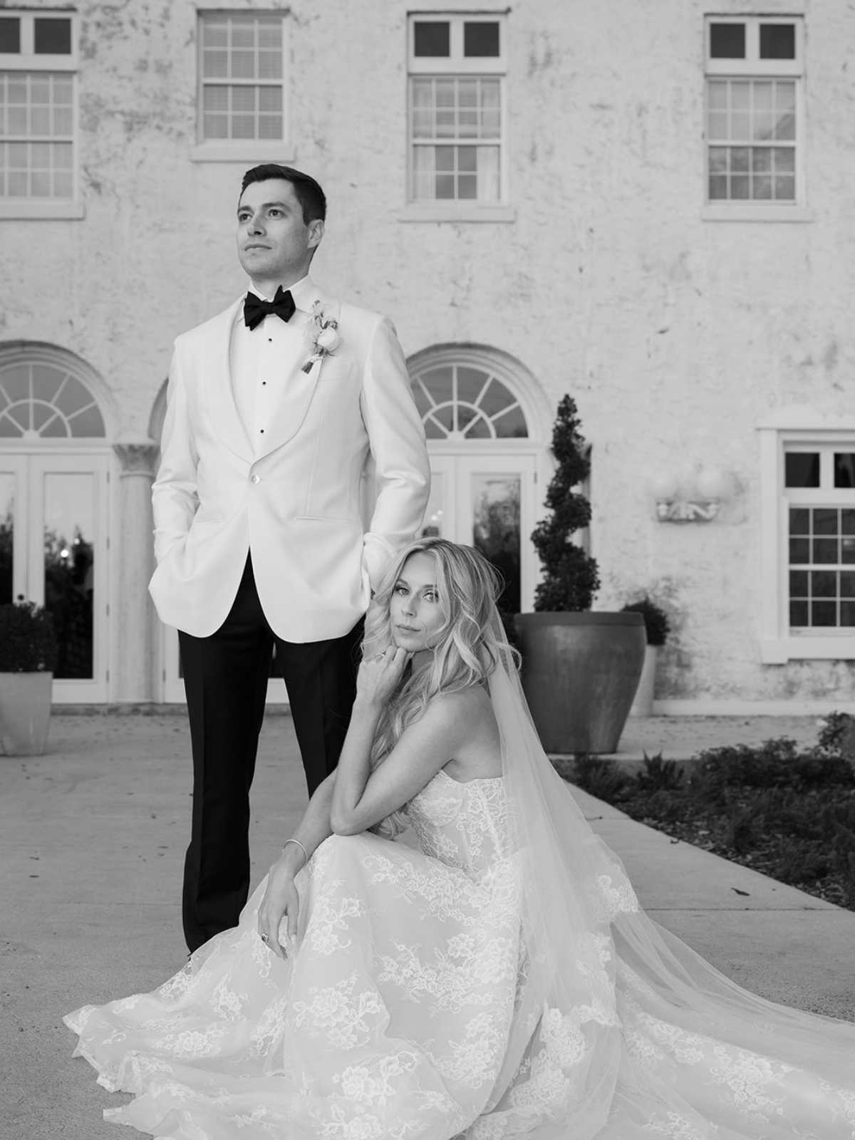 Black and white photo of a bride in a wedding dress sitting on the ground with a long veil, and a groom in a tuxedo with a white jacket and black bow tie standing behind her with hands in pockets, in front of a building with arched windows and potted plants.
