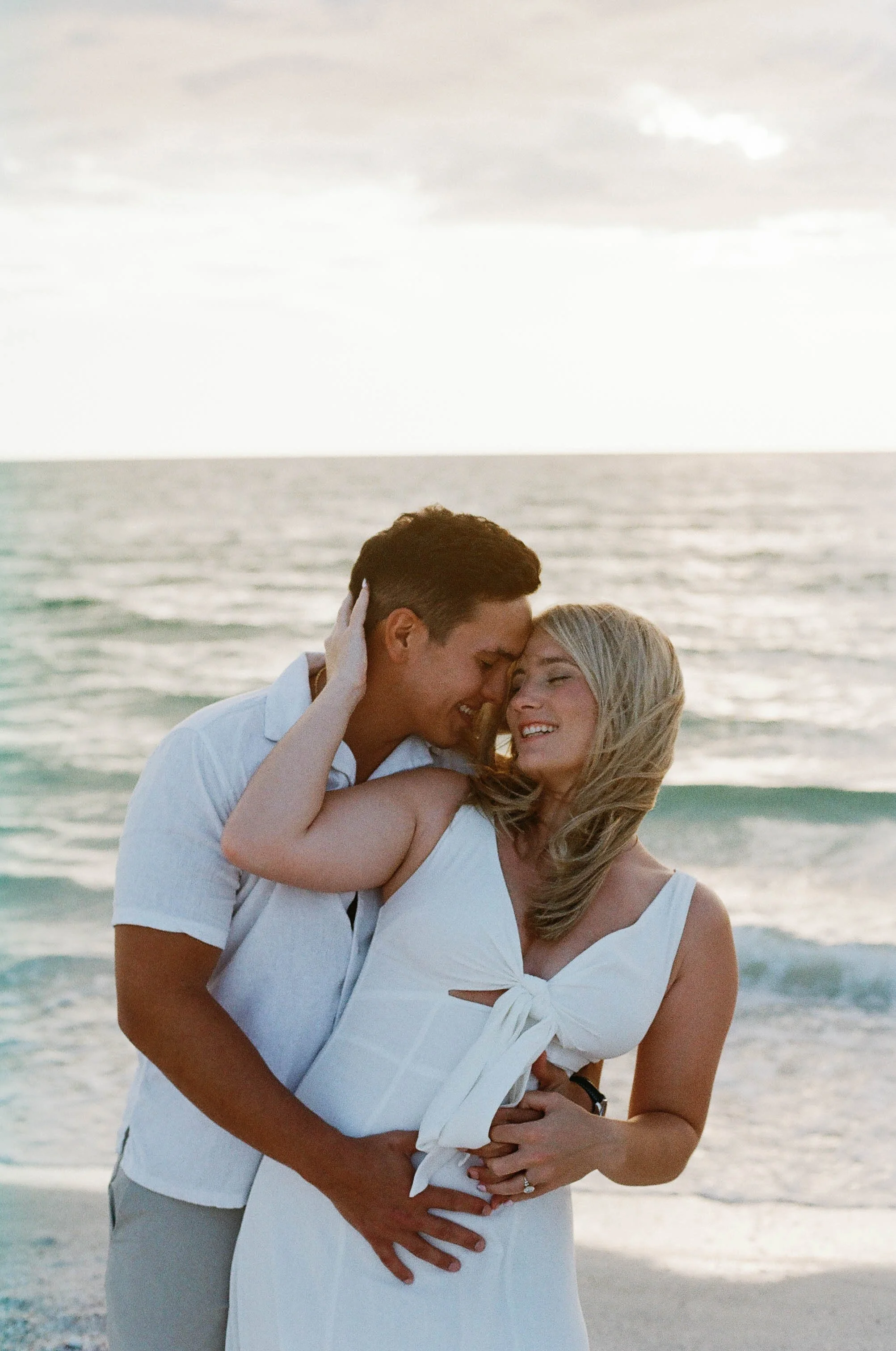 A couple embracing at the beach. The man is gently holding the woman's face and they are touching foreheads, smiling with closed eyes, in a romantic moment.