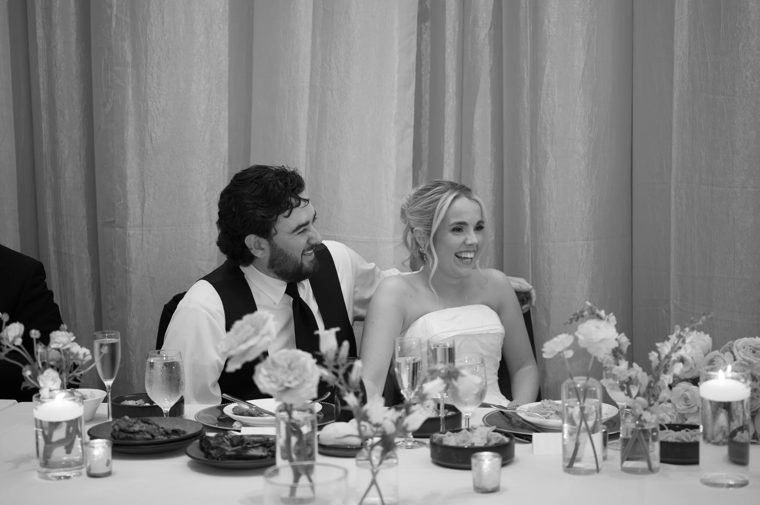 A black and white photo of a bride and groom sitting at their wedding reception table, smiling and enjoying the moment. The table is decorated with flowers and arranged with various dishes and drinks.