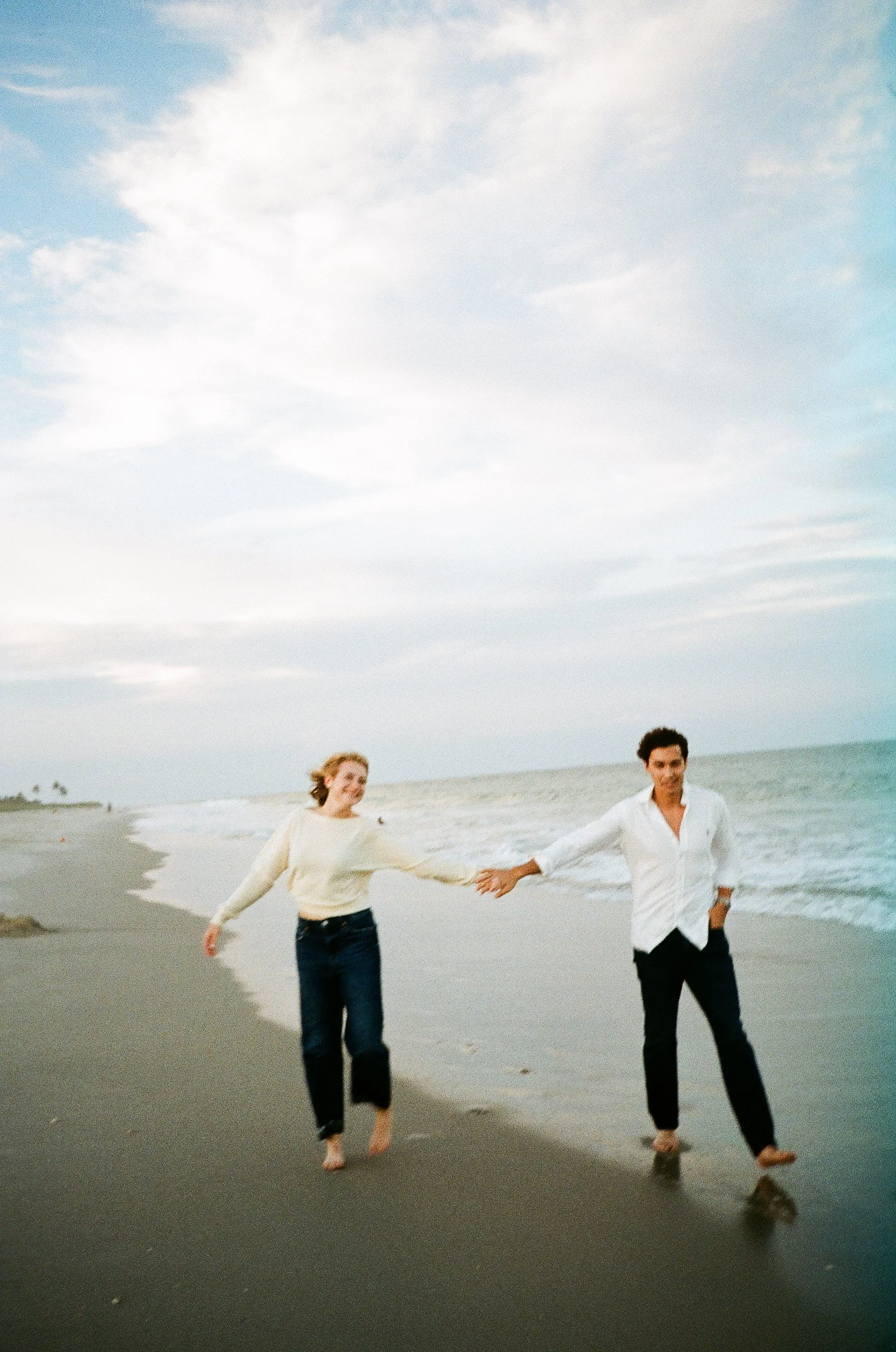 A man and a woman holding hands and walking barefoot along the shoreline at the beach, with a partly cloudy sky overhead.