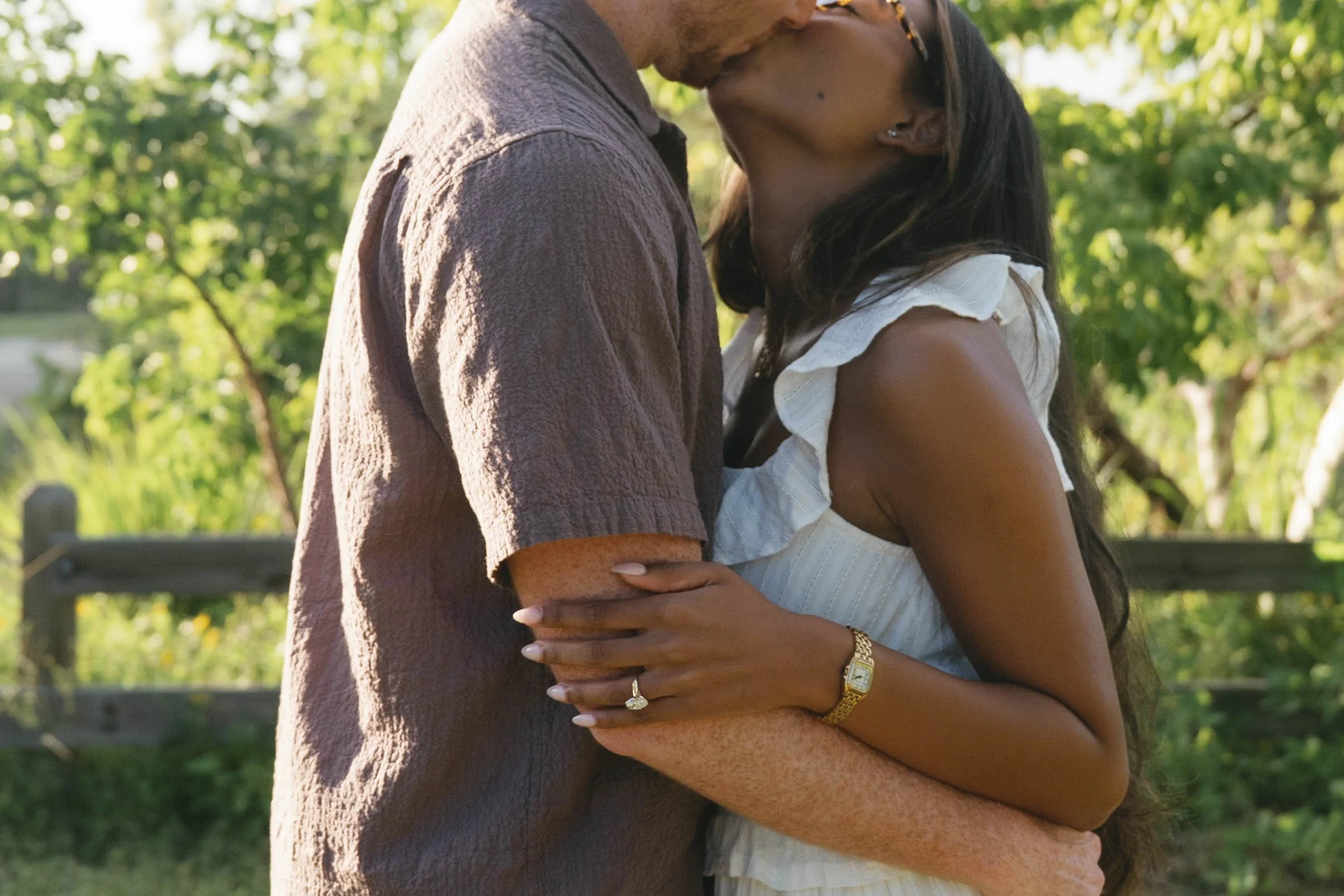 A couple sharing an intimate moment, embracing with foreheads touching outdoors in a park with green trees in the background.