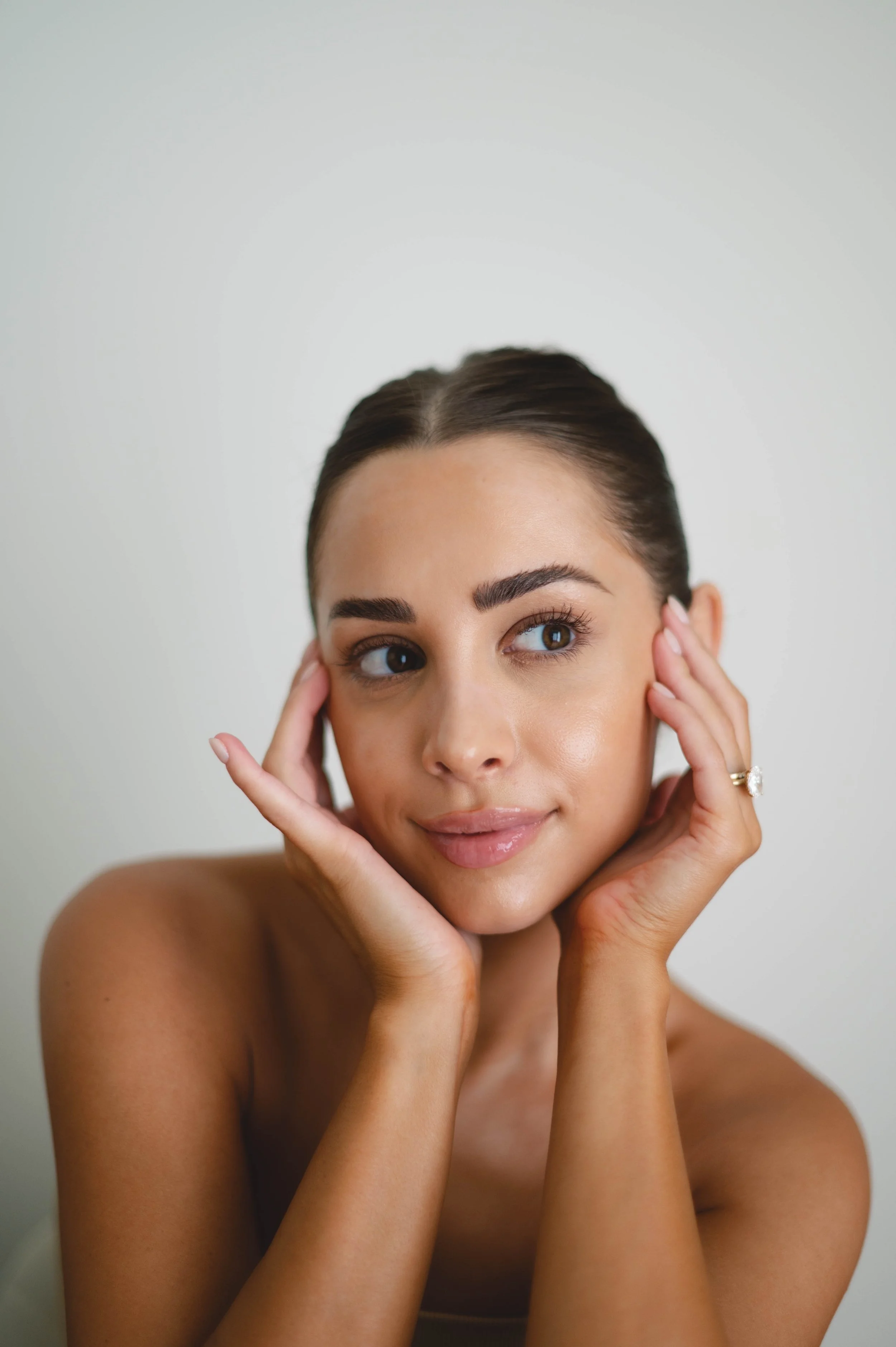 Close-up of a young woman with brown hair, brown eyes, and light makeup, posing with her hands gently touching her face against a plain backdrop.