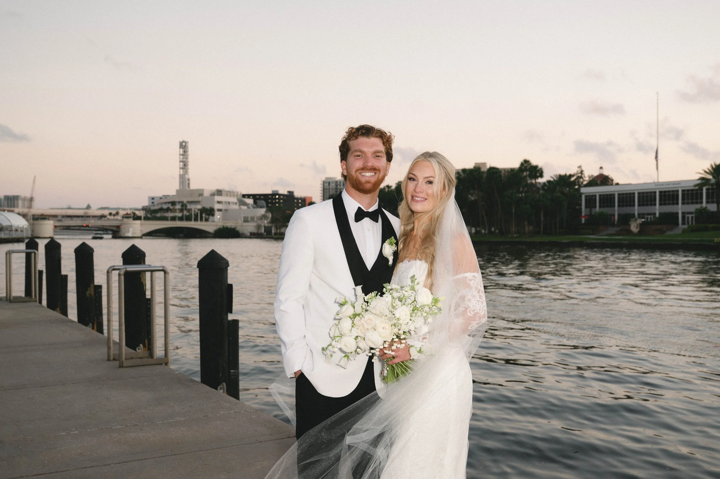 A newlywed couple in wedding attire posing by a river at sunset, with city buildings and trees in the background.