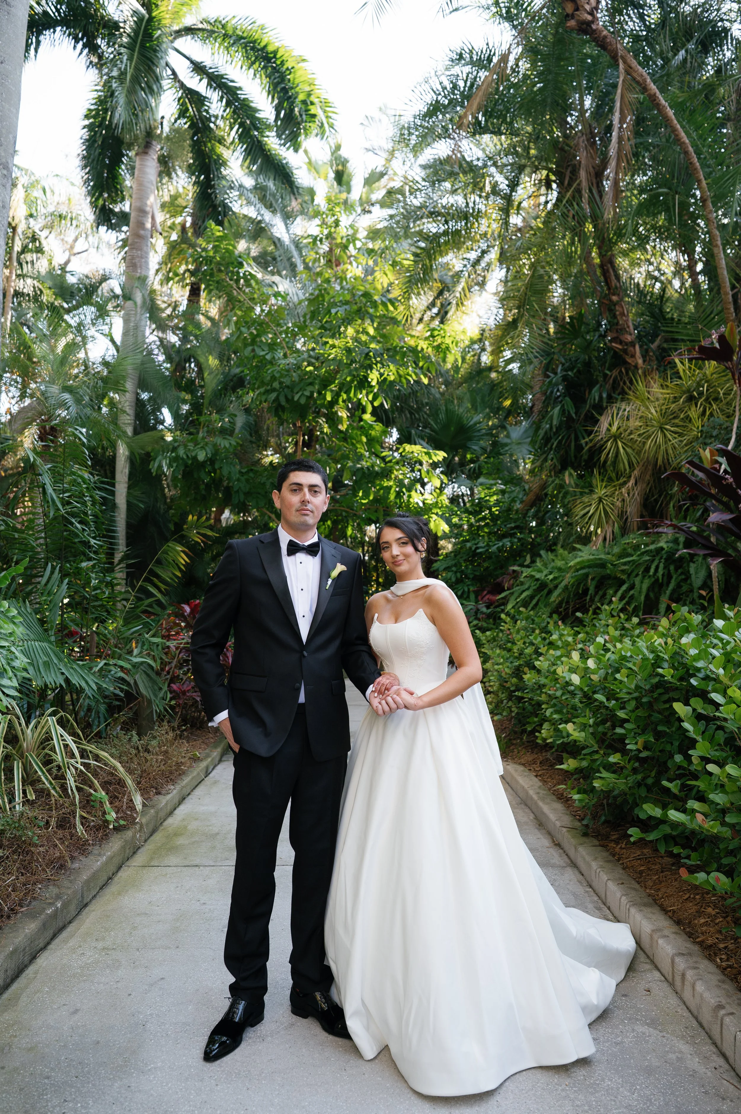 A bride and groom stand together on a garden pathway surrounded by lush green tropical plants. The groom wears a black tuxedo with a bow tie, and the bride wears a white strapless wedding gown with a sweetheart neckline, holding a bouquet. They are p