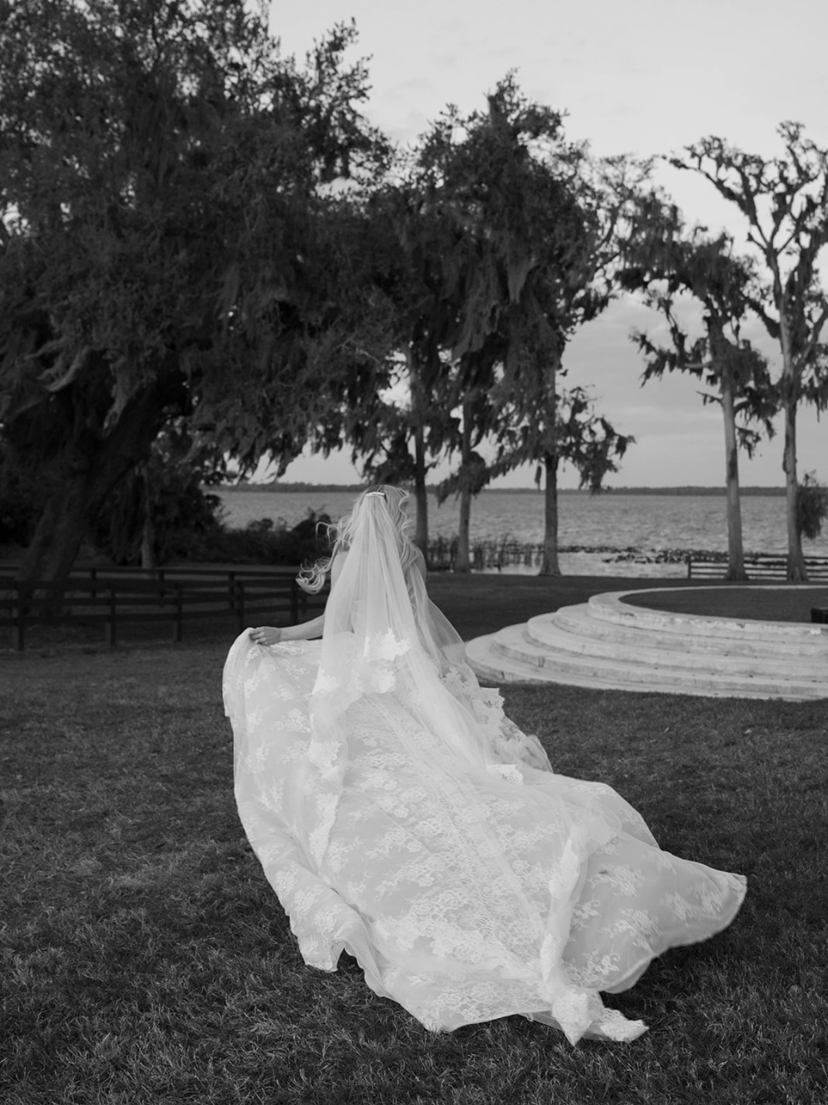 A woman in a wedding dress with a veil walks in a park near a lake, with trees and benches in the background, in black and white.