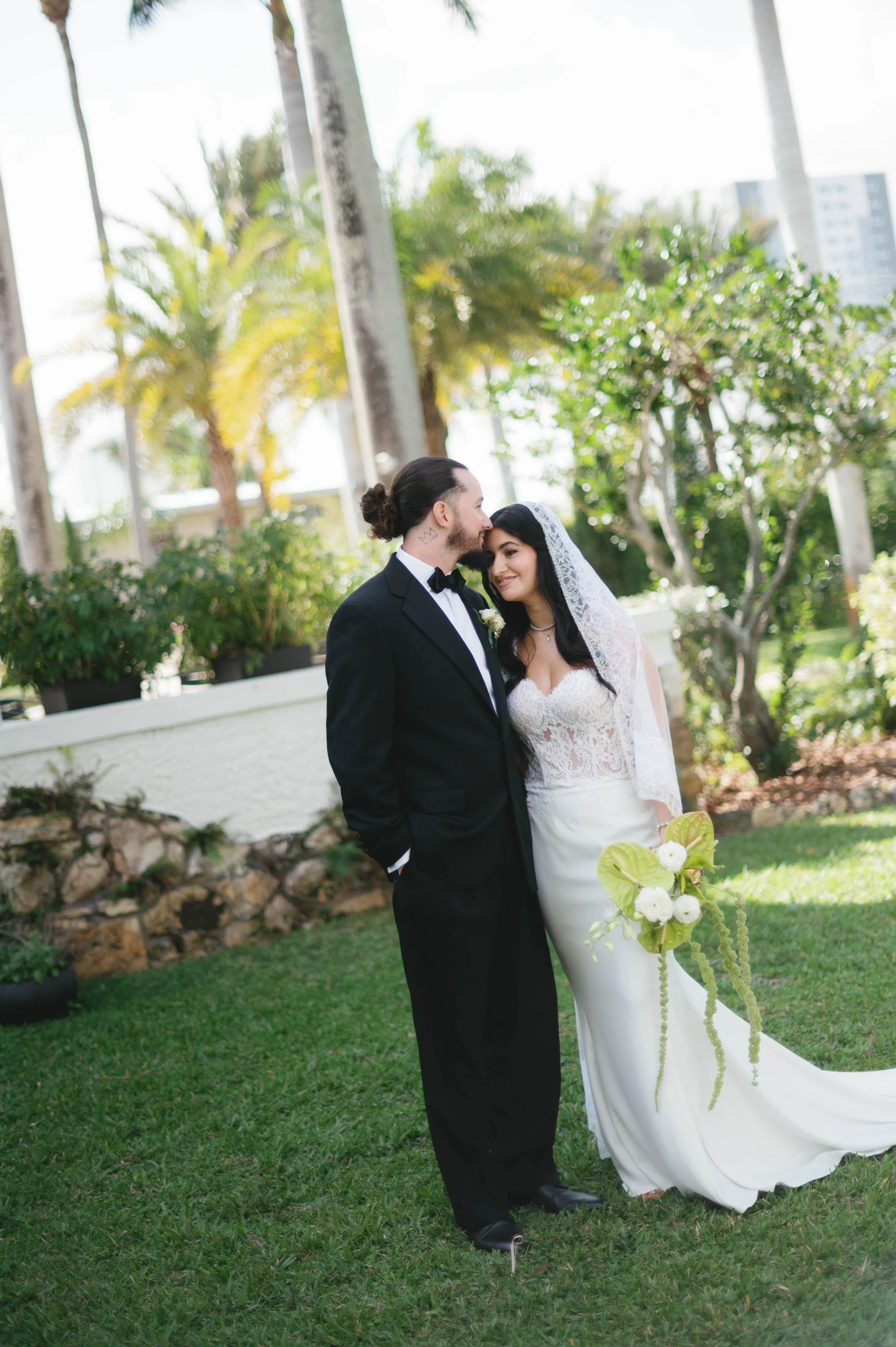 A bride and groom standing outdoors in a garden, sharing an intimate moment. The groom, in a black tuxedo, is kissing the bride on her forehead. The bride is in a white wedding gown with a lace veil, holding a bouquet of white flowers and greenery. P