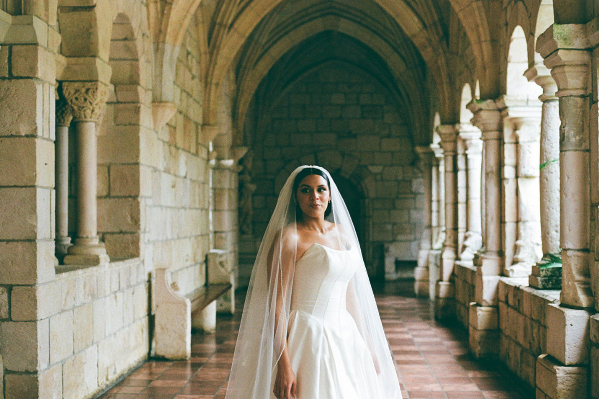 Bride in a white wedding dress and veil, standing in a stone arched corridor with columns and checkered floor.