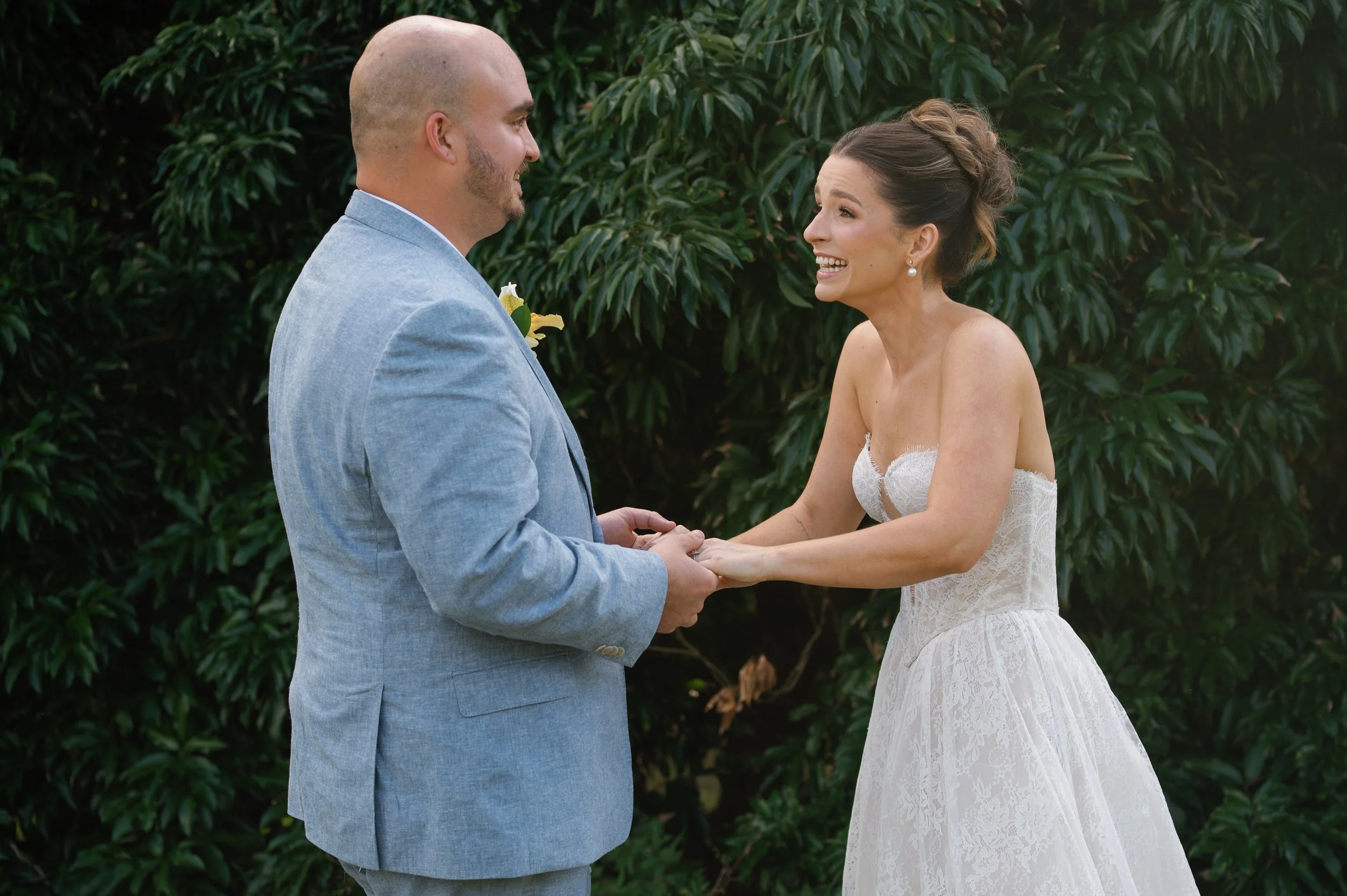 A bride and groom exchanging vows outdoors, holding hands, with green foliage in the background.