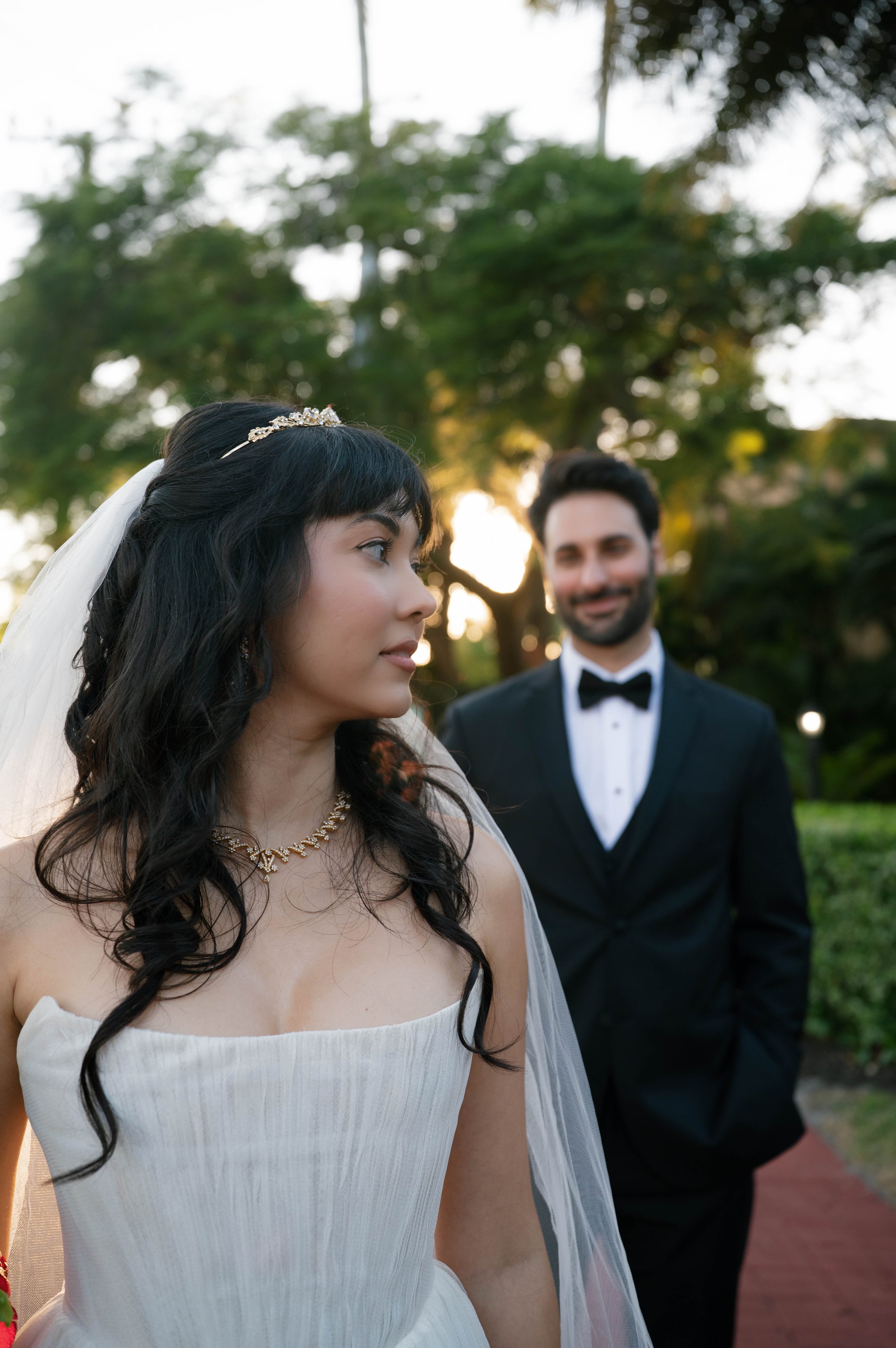 A bride with long dark hair and a tiara, wearing a white gown with thin straps, looking to the side, with a groom in a black tuxedo and bow tie looking at her in the background, set outdoors during sunset with trees and greenery.