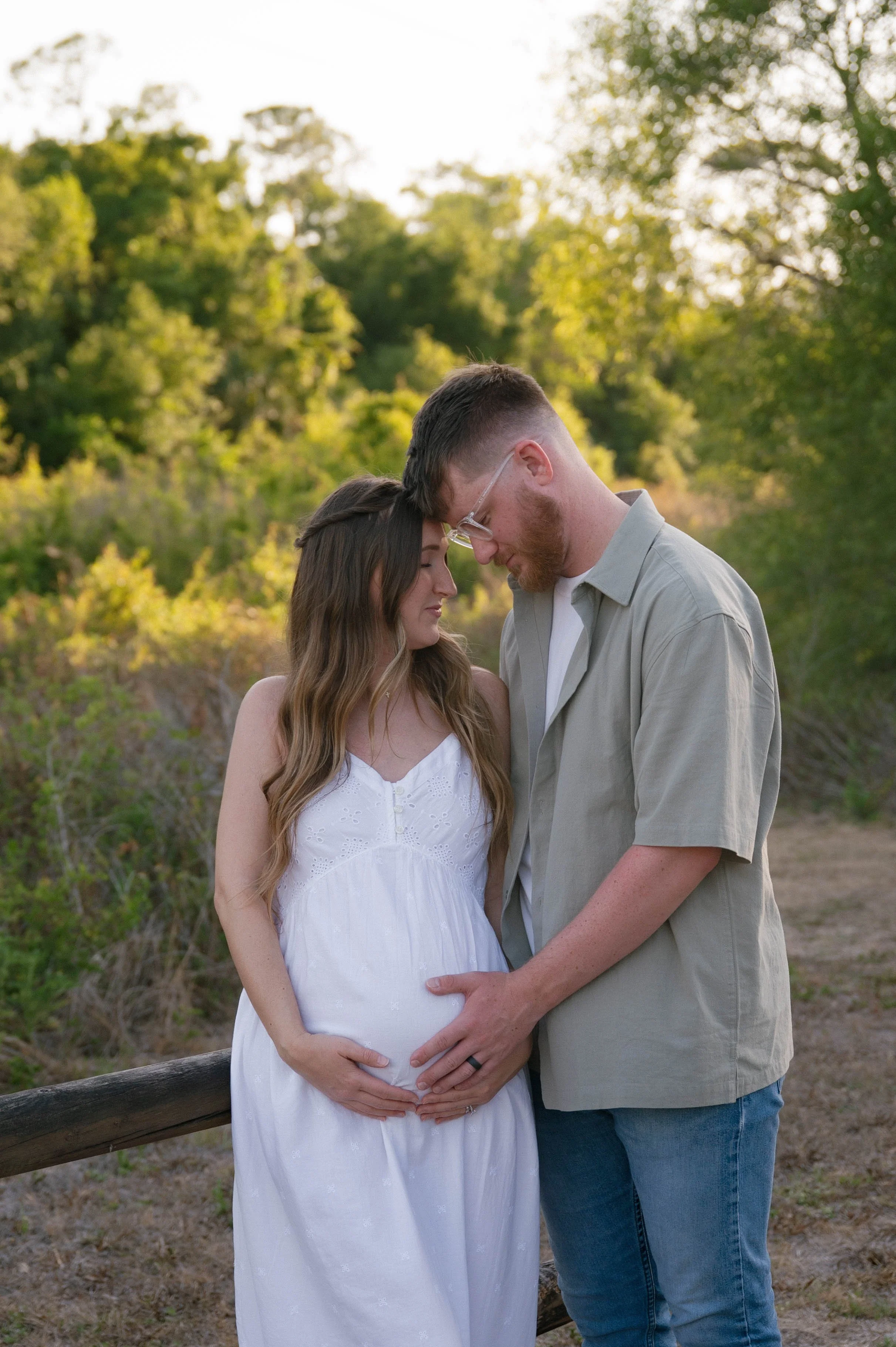 A pregnant woman in a white dress and a man in glasses and a light gray shirt share a moment, touching her belly and foreheads, outdoors with green trees in the background during golden hour.
